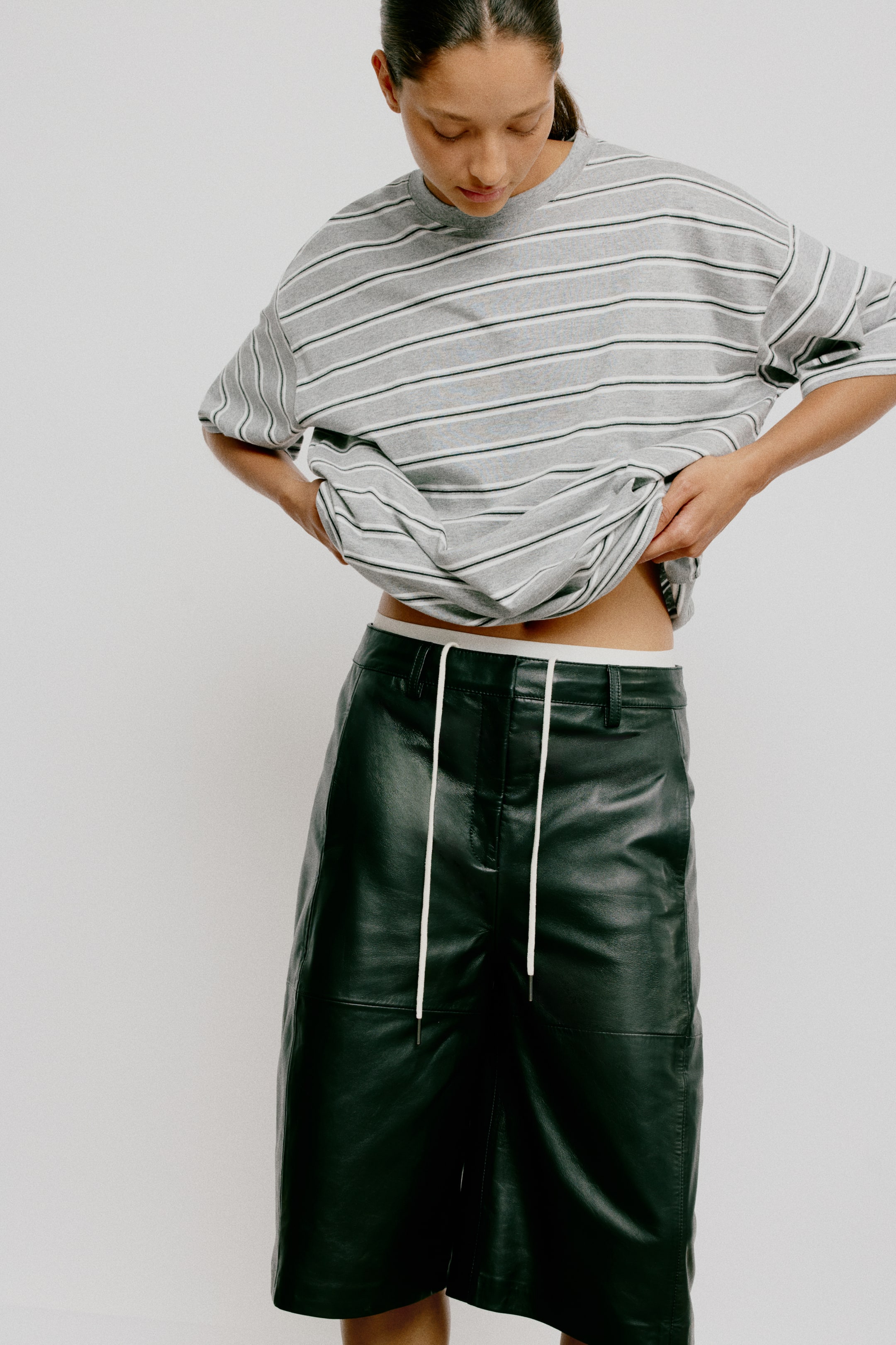 Ver imagen más grande: Una camiseta extragrande en color gris, estampada con rayas horizontales blancas y negras, está siendo llevada por la modelo. Presenta un ribete acanalado alrededor del cuello y hombros caídos, y su dobladillo de corte recto es levantado por las manos de la modelo, revelando el abdomen y la parte superior de unos pantalones cortos de cuero verde oscuro.