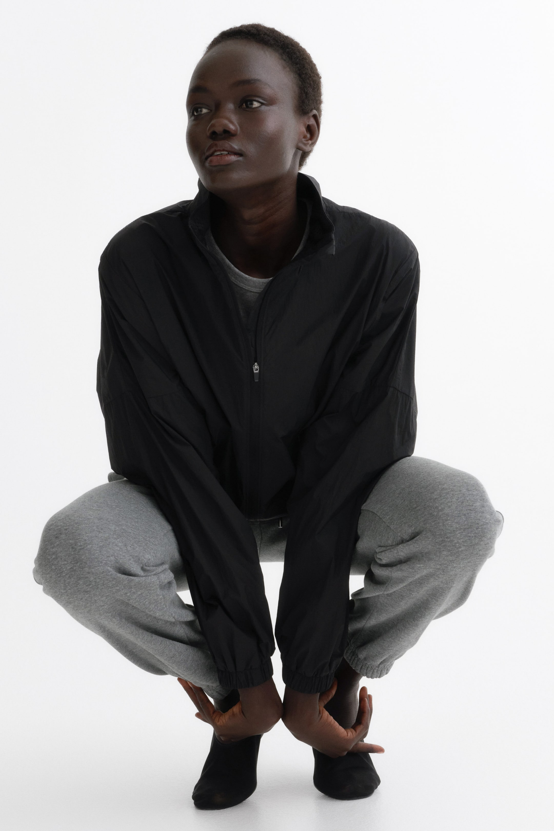 Voir une image plus grande: Une veste légère noire est présentée de face, avec un col montant, une fermeture éclair frontale complète de ton argenté et de longues manches dotées de poignets et d’un ourlet élastiqués, portée par-dessus un t-shirt gris.