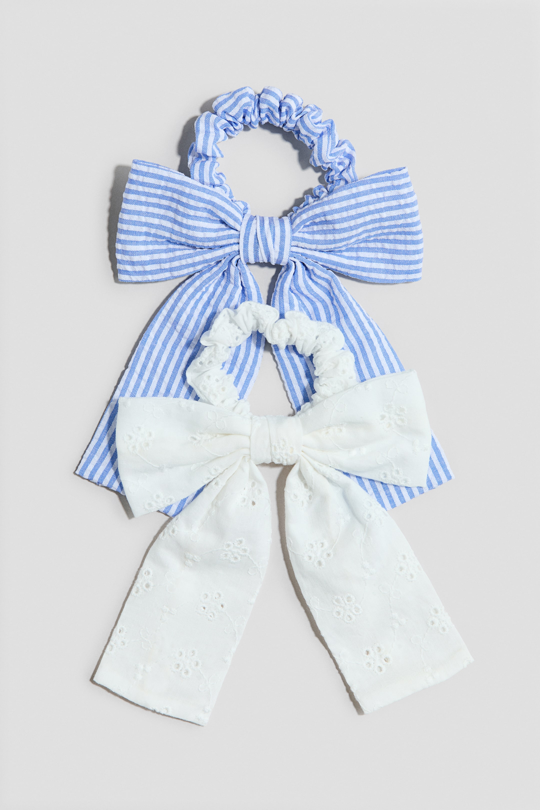 View larger image: Two hair scrunchies are featuring a ruffled elastic band and a large bow with long tails. One scrunchie is blue and white striped, while the other is white with eyelet embroidery, layering over the striped scrunchie.