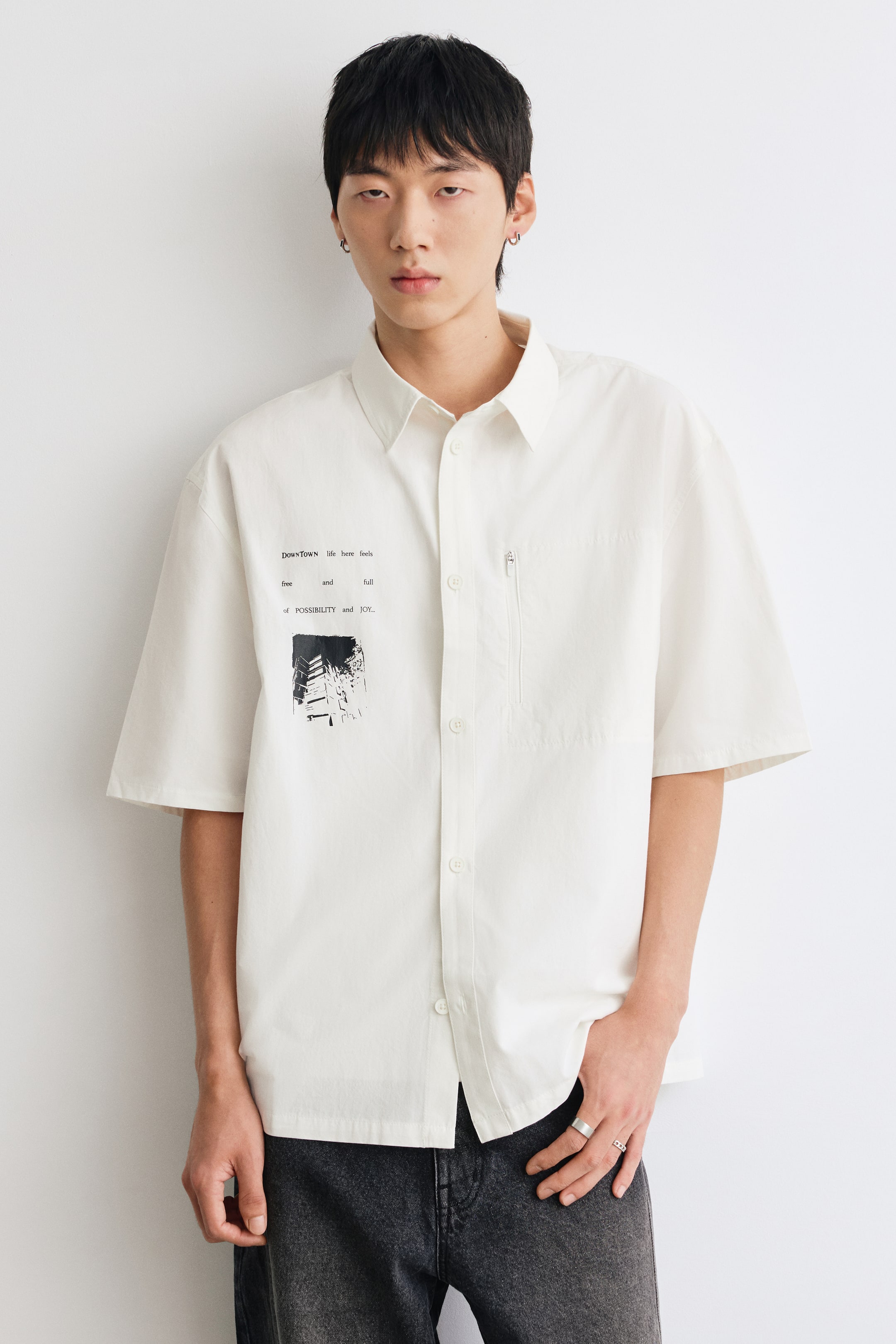 Xem ảnh lớn hơn: Người mẫu đang mặc một chiếc áo sơ mi cotton tay ngắn màu trắng ngà, phom rộng, cổ điển và cài nút phía trước. Áo được nhấn bằng họa tiết đồ họa và chữ màu đen ở ngực phải cùng một túi khóa kéo dọc ở bên trái, phối với quần jeans xám đậm.