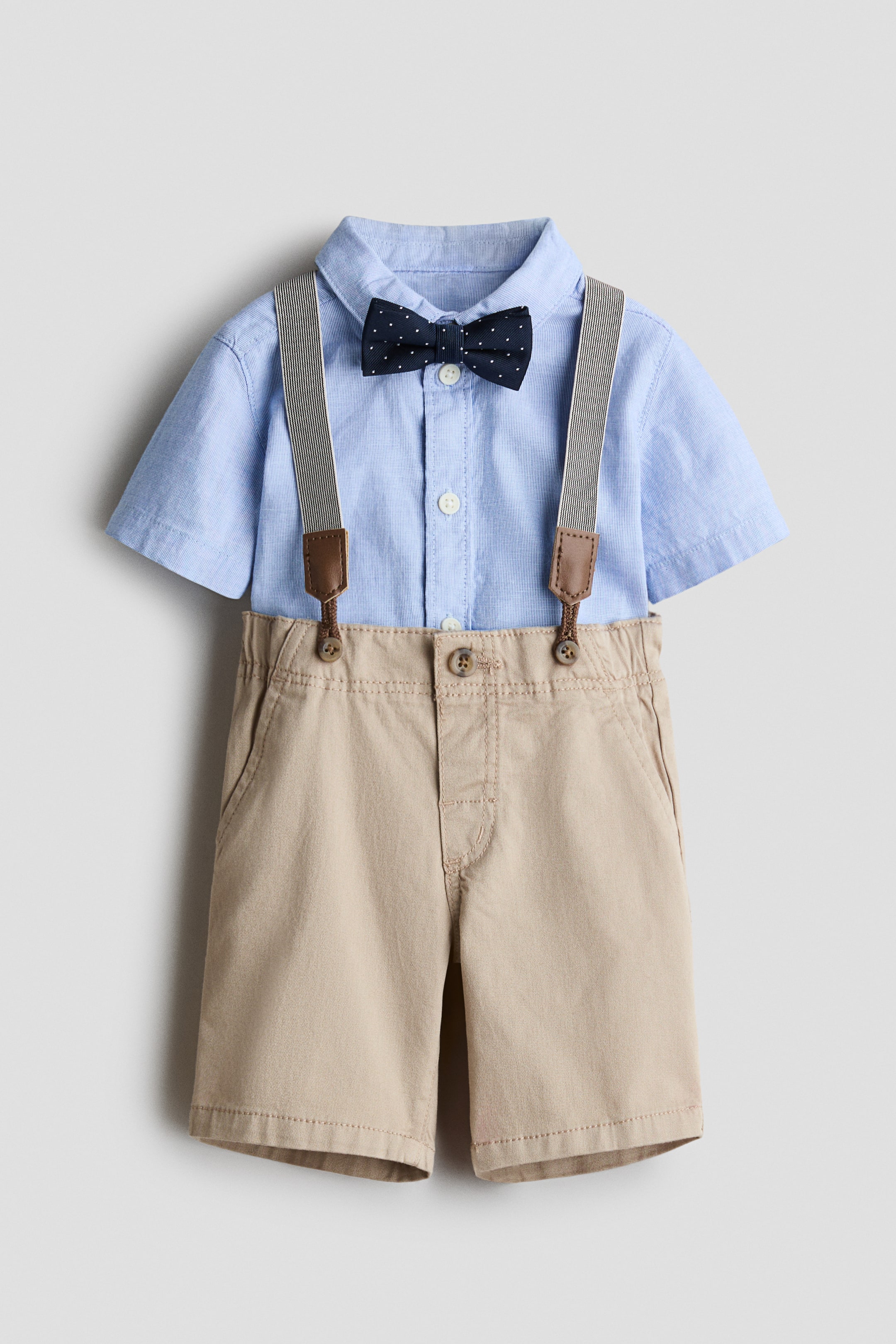 View larger image: A sophisticated ensemble is showcased from the front, featuring a light blue, short-sleeved shirt with a button-down collar and white buttons, accompanied by a dark blue polka dot bow tie at the collar. The shirt is paired with khaki shorts, which include detachable striped braces with brown accents, side pockets, and a faux fly with a button.