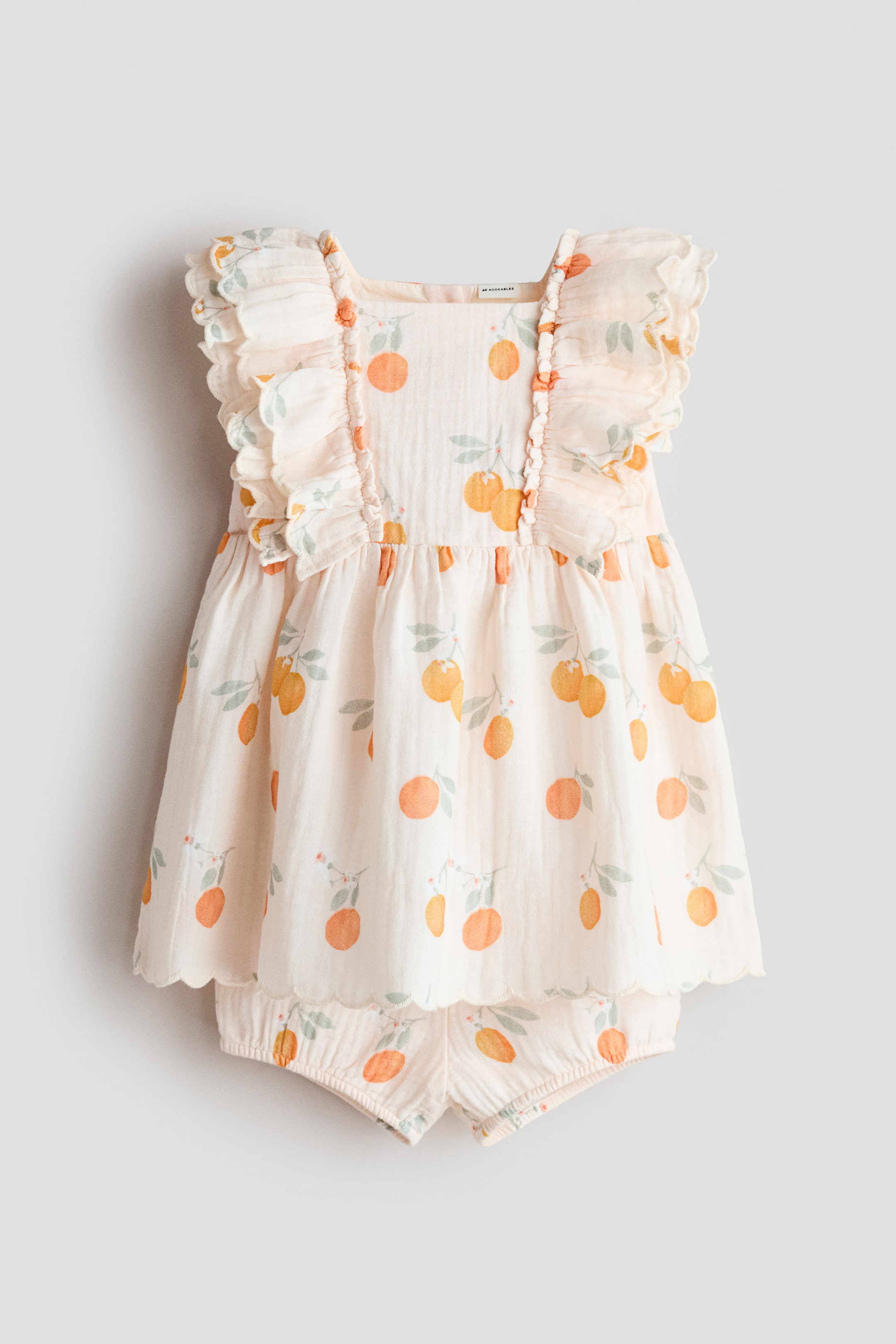 View larger image: The H&M Adorables dress and bloomers set, facing front, features a light-coloured muslin fabric with an orange fruit and leaf pattern. The dress showcases a square neckline with a narrow-cut yoke adorned with double-layered, scalloped frills continuing over the shoulders, gathers at the waist, and a flared skirt with a scalloped hem, while the matching bloomers display elastication at the leg openings.