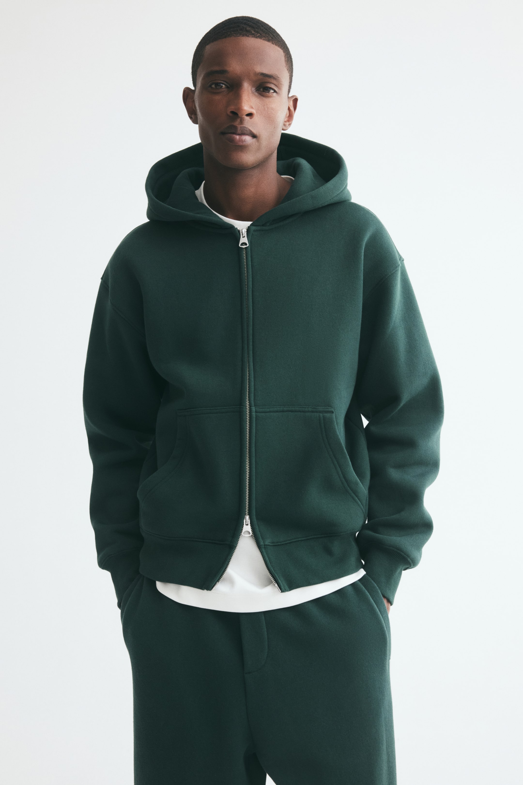 View larger image: The model is wearing a dark green zip-up hoodie, layered over a white t-shirt and styled with matching sweatpants. The relaxed-fit hoodie features a hood, dropped shoulders, front pockets, ribbed trims, and a silver two-way zipper.