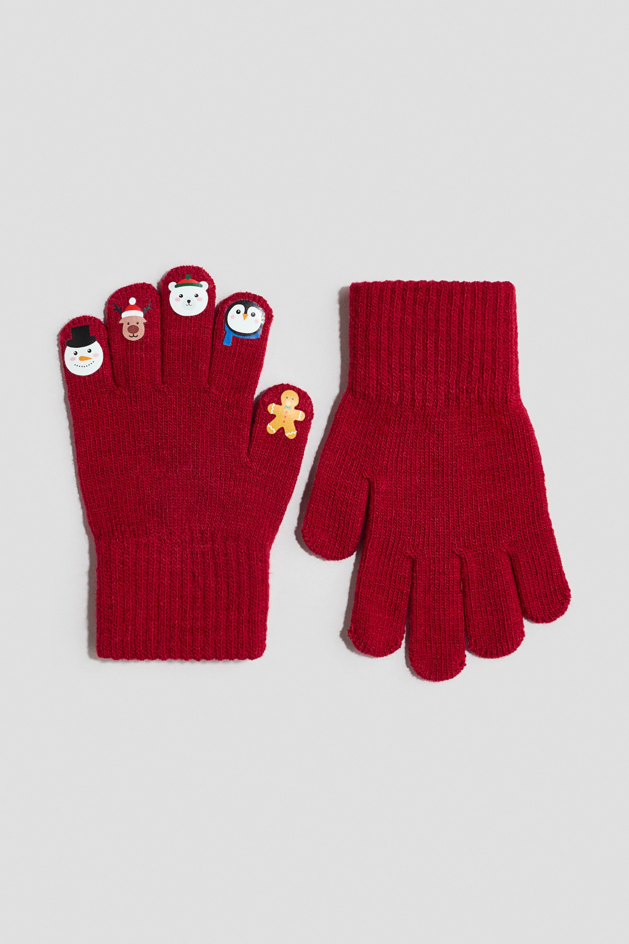 Größeres Bild ansehen: Ein Paar rote Feinstrickhandschuhe wird mit Vorder- und Rückansicht gezeigt und verfügt über breite Rippbündchen. Der nach vorne gerichtete Handschuh präsentiert an den Fingerspitzen Cartoon-Motive, darunter einen Schneemann, ein Rentier, einen Eisbären, einen Pinguin und ein Lebkuchenmännchen, die das touchscreen-kompatible Material darstellen.