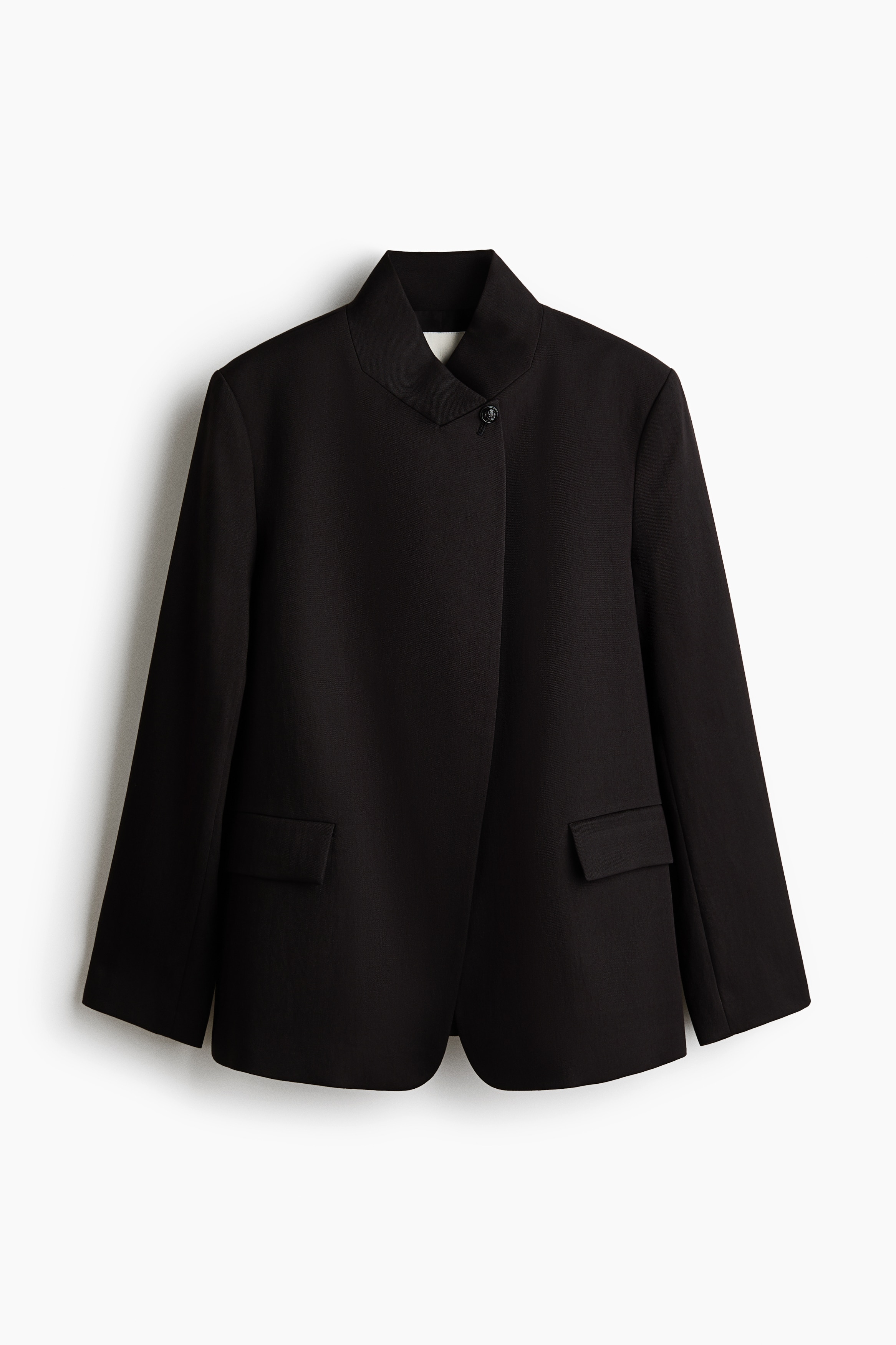 The model is wearing an oversized, dark brown woven blazer featuring a high stand collar, structured shoulders, and a wrap-front closure with a single visible button at the neck. The blazer is styled with slim black trousers, long black leather gloves, and a black clutch bag tucked under the arm.