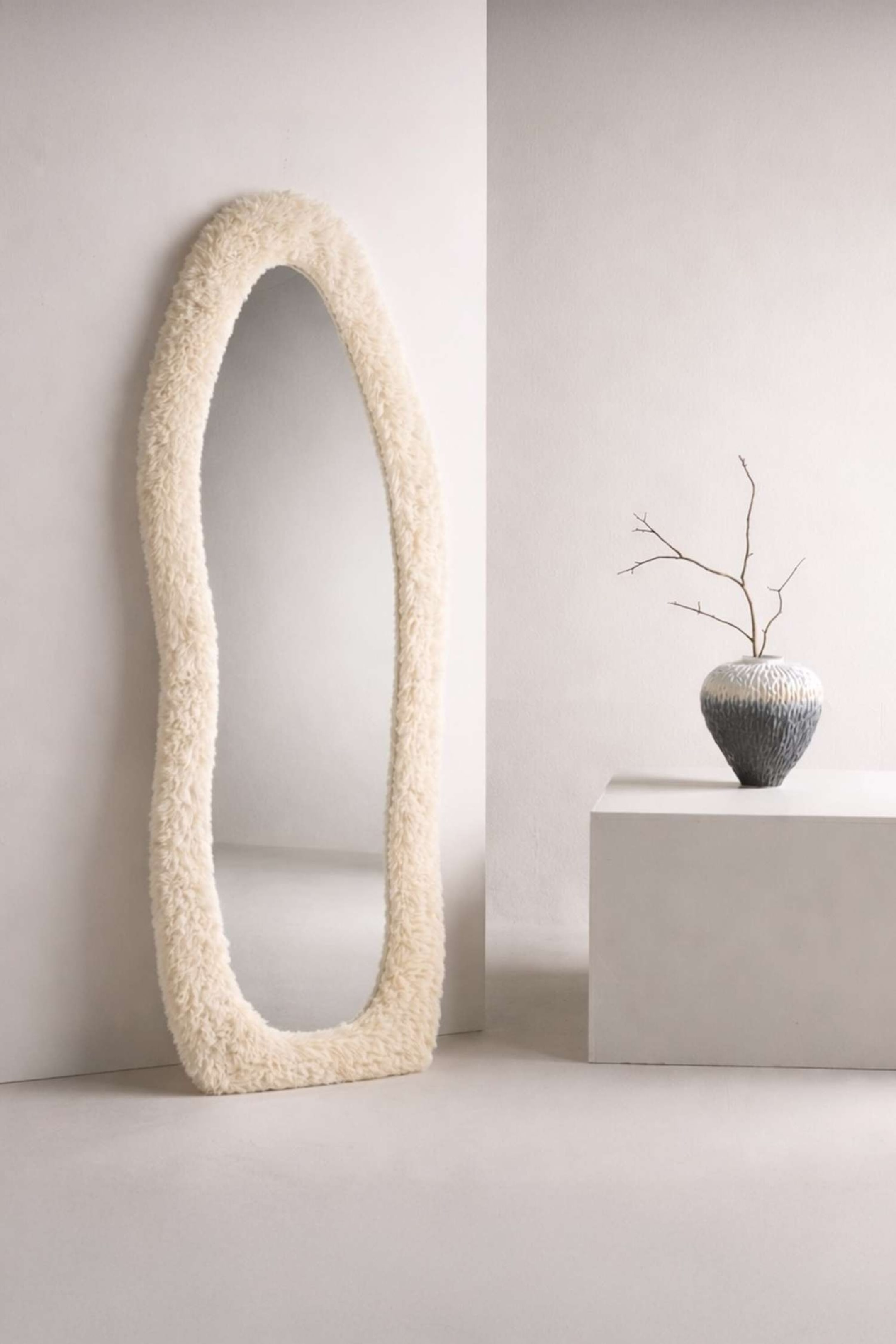 Visualizza immagine più grande: Uno specchio a figura intera è posizionato verticalmente, caratterizzato da un contorno irregolare e ondulato. È incorniciato con un materiale spesso in finta pelliccia color crema, che presenta un aspetto morbido e strutturato.