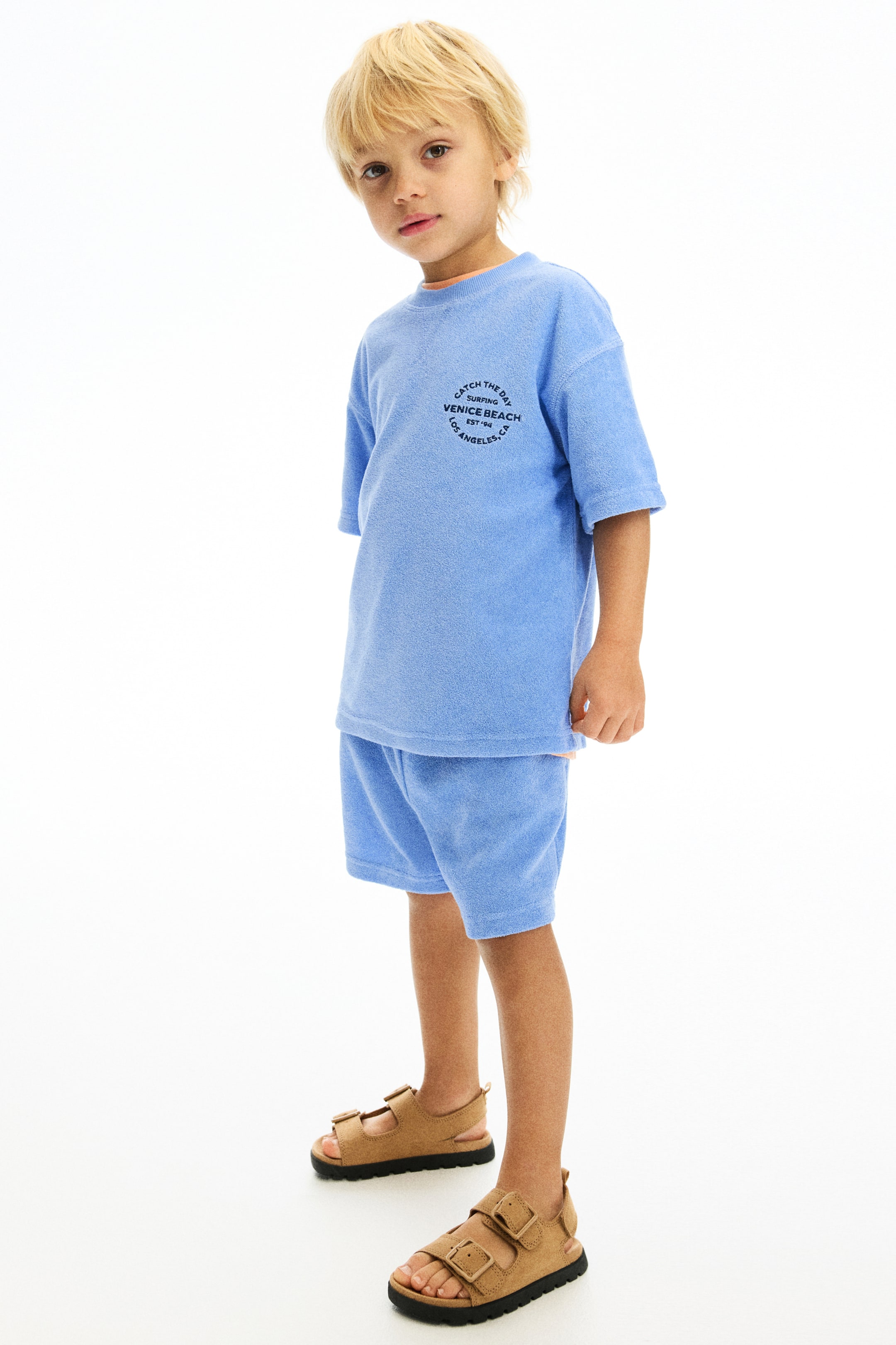 Prikaži veću sliku: Plavi set od frotira koji se sastoji od majice kratkih rukava i odgovarajućih šortseva prikazan je s prednje strane. Majica ima kružni crni grafički print na grudima, a šortsevi dosežu do sredine butine. Uz set se nose smeđe sandale sa dve kaiševe i kopčama.