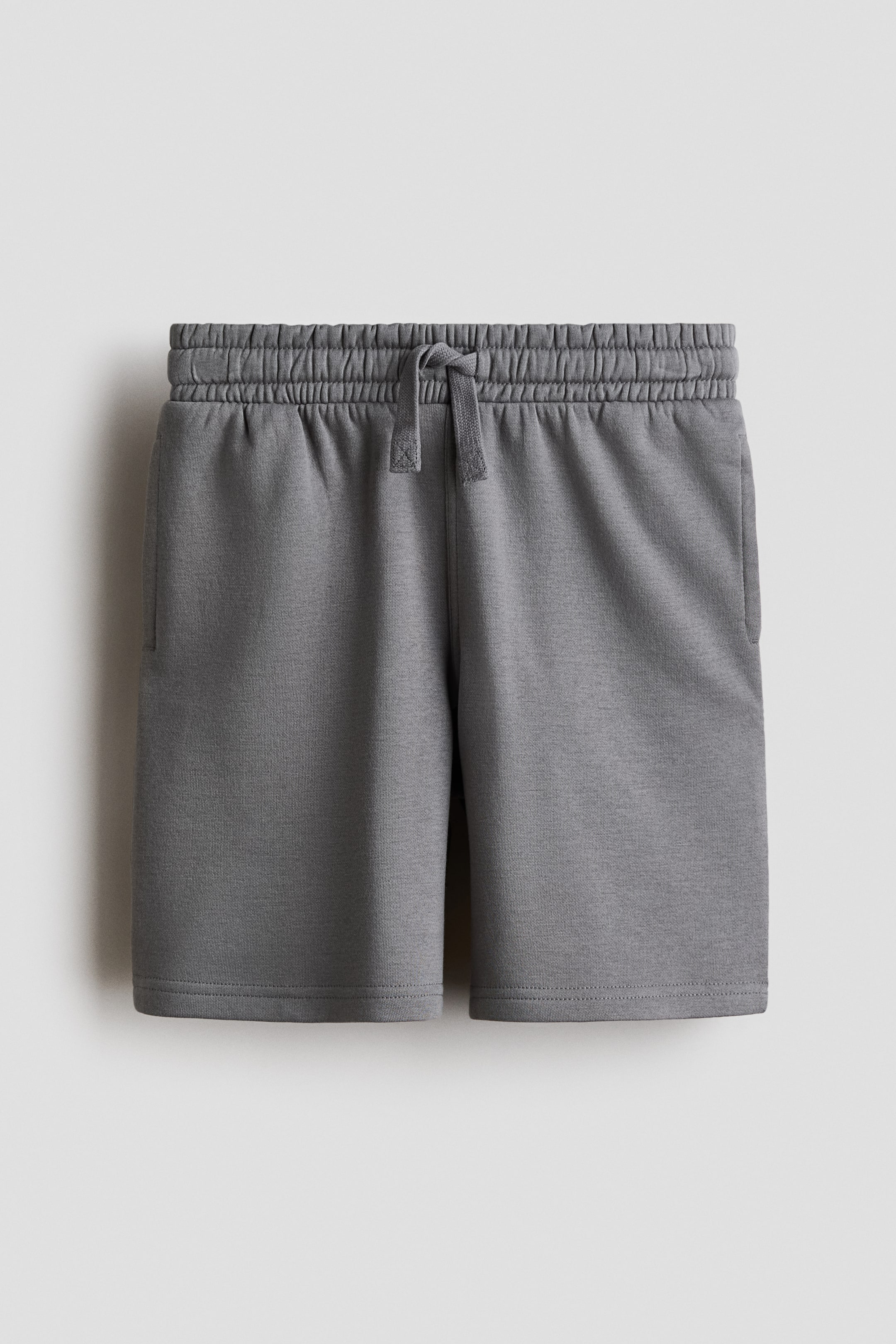 Ver imagen más grande: Pantalones cortos grises a la altura de la rodilla se presentan desde el frente, confeccionados con una tela de sudadera gris jaspeada. Cuentan con una cintura elástica con cordón ajustable, el cual está atado, y bolsillos discretos tipo ojal son visibles en las costuras laterales posicionadas al frente.