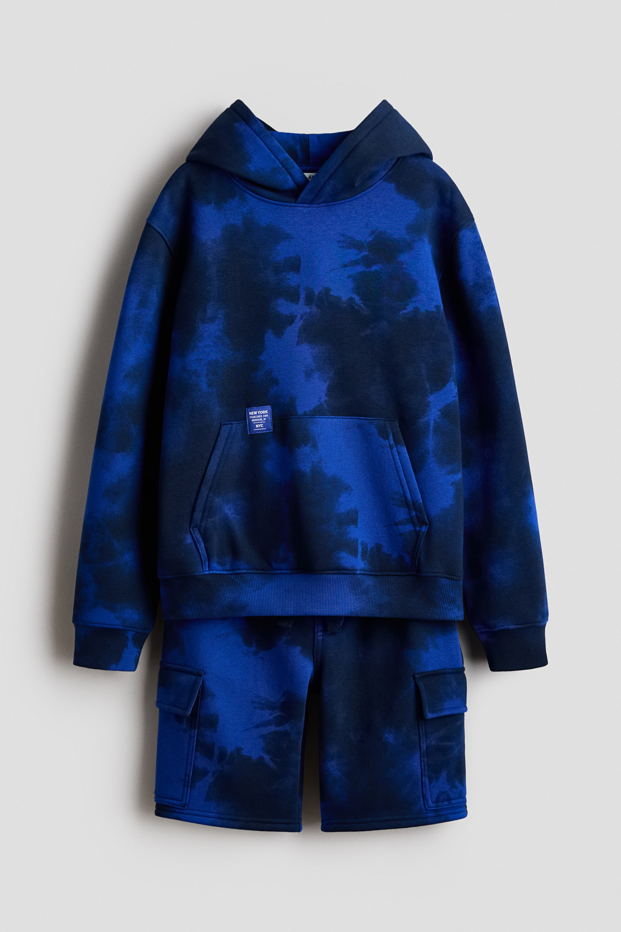 Visa större bild: Ett blått tie-dye set med huvtröja och shorts visas framifrån. Huvtröjan har en huva med jerseyfoder, nedhasade axlar, långa ärmar med ribbade muddar, en ribbad nederkant och en känguruficka. De matchande shortsen har en elastisk midja och fickor med lock på benen.