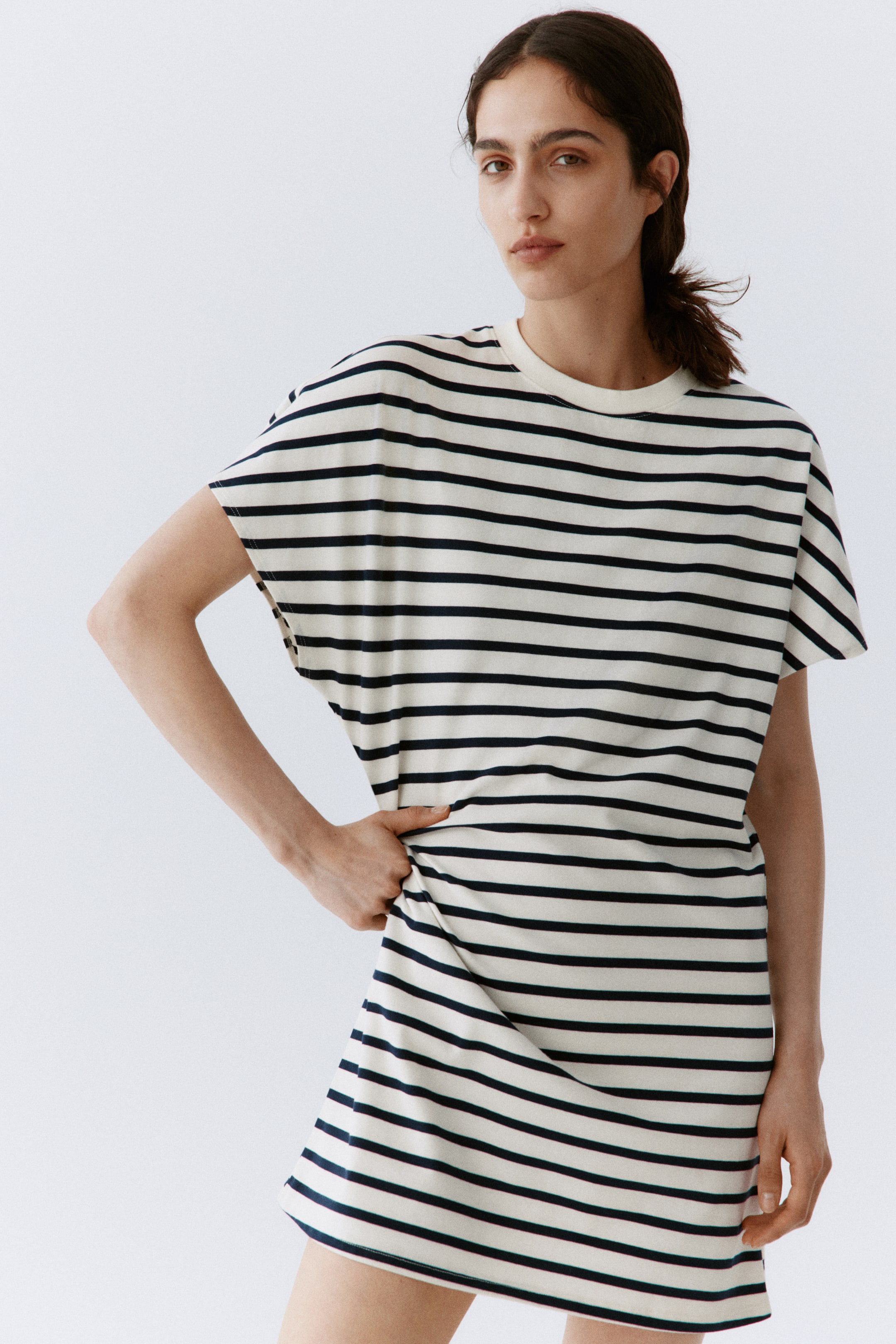 Agrandir l'image: Une robe t-shirt à rayures présente une base blanc cassé avec des rayures horizontales noires, un col rond et des manches courtes amples. Elle tombe à mi-cuisse avec une coupe décontractée, légèrement inclinée vers la droite, et son tissu est doucement tiré à la hanche, mettant en valeur son drapé.