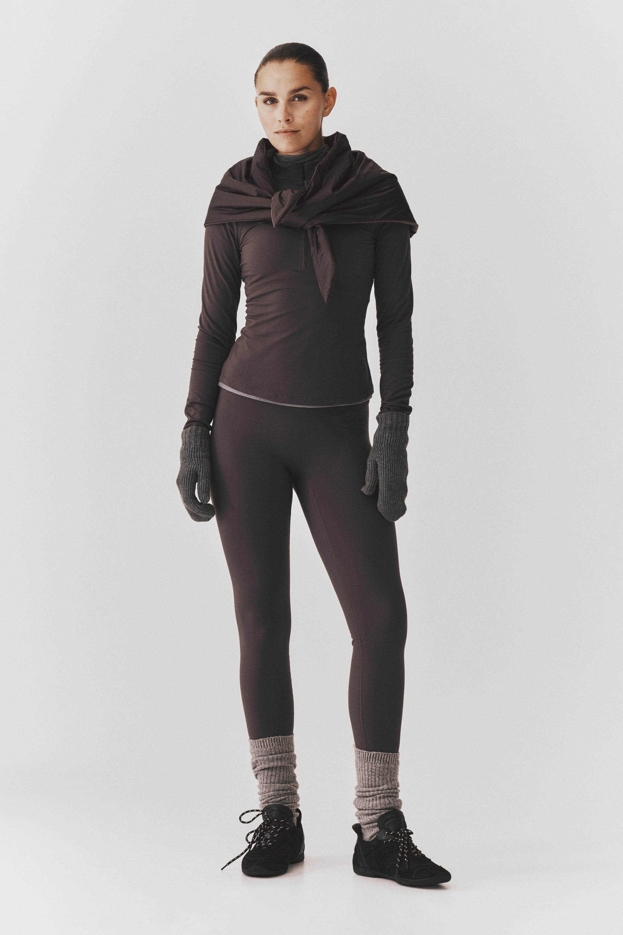 Voir une image plus grande: Un ensemble de vêtements de sport brun foncé composé d’un haut ajusté à manches longues et de leggings assortis est porté par le modèle. Une veste coordonnée est nouée autour des épaules, tandis que des mitaines en tricot grises, des jambières côtelées et des chaussures de sport noires à lacets complètent la tenue.