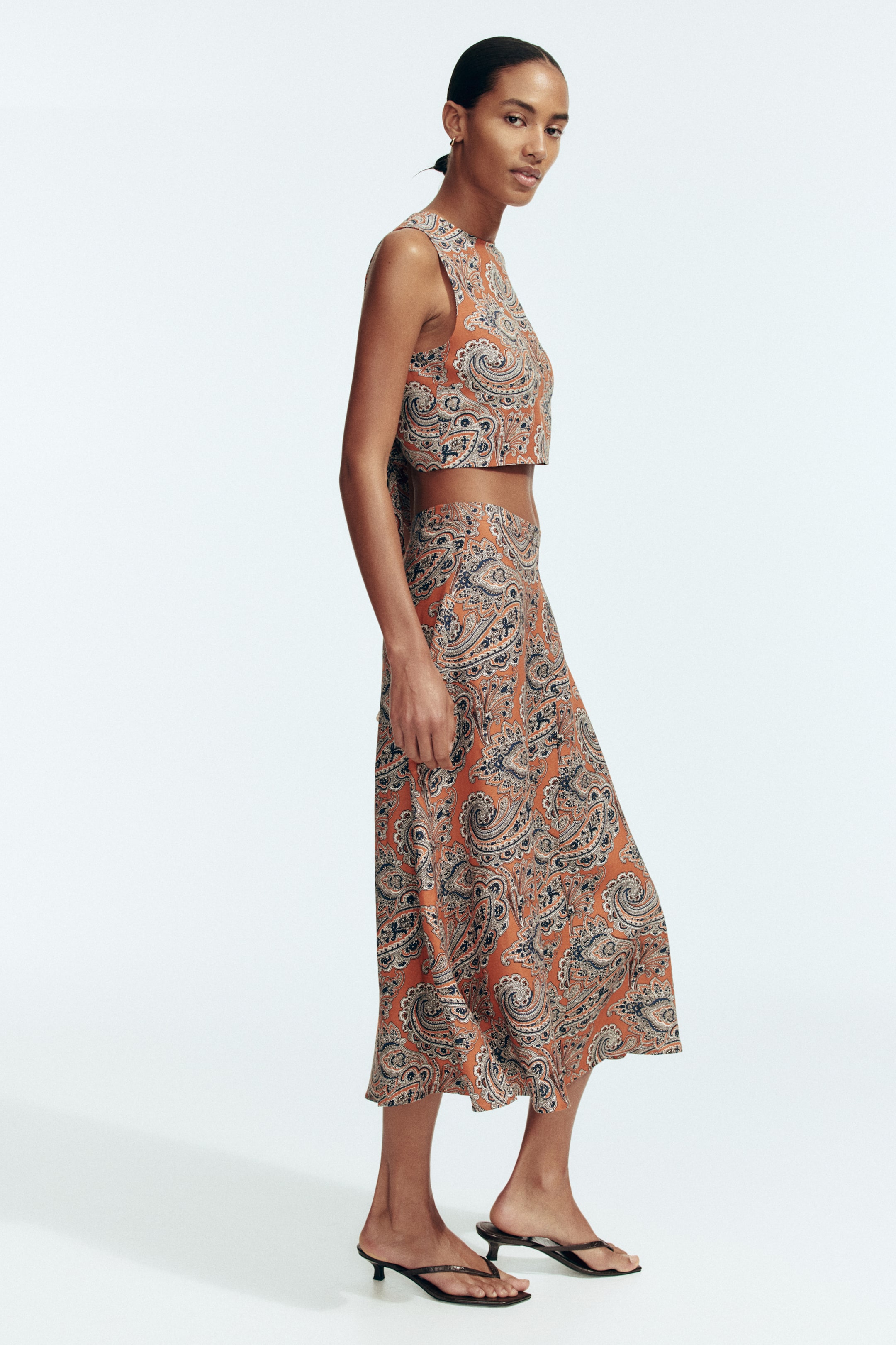 View larger image: A two-piece set, featuring a sleeveless crop top and a flowing midi skirt, is displaying a rust orange, black, and white paisley print. The top features a high neckline and a cut-out detail at the back, while the skirt falls to mid-calf.