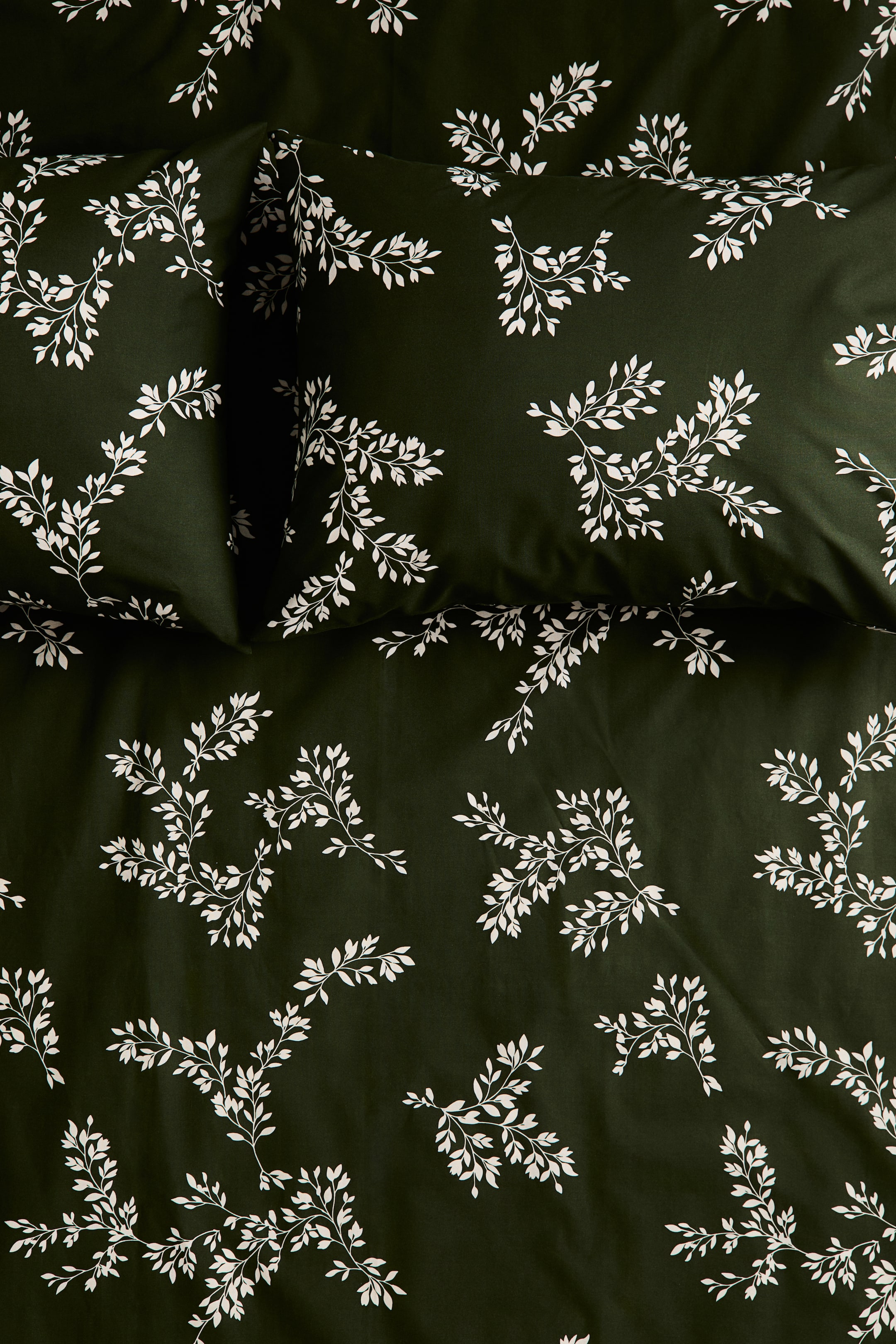 View larger image: A dark green bedding set, including a duvet cover and matching pillowcases, is showcasing a repeating pattern of delicate white botanical leafy sprigs in an overhead view. The smooth fabric is arranged to display the coordinated design across both the pillows and the main cover.