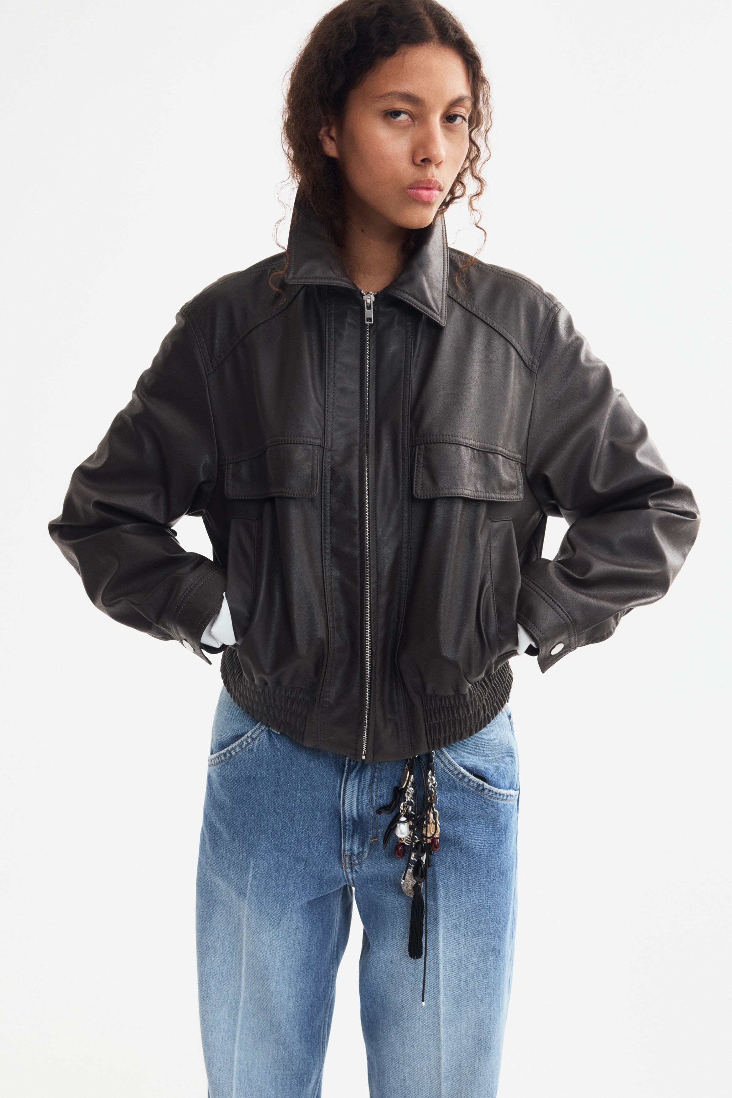 Une veste en cuir marron foncé est présentée de face, avec un col à revers et une fermeture éclair argentée descendant au centre. Deux grandes poches à rabat sont visibles sur le bas, et la veste est dotée d'une large ceinture élastique smockée.