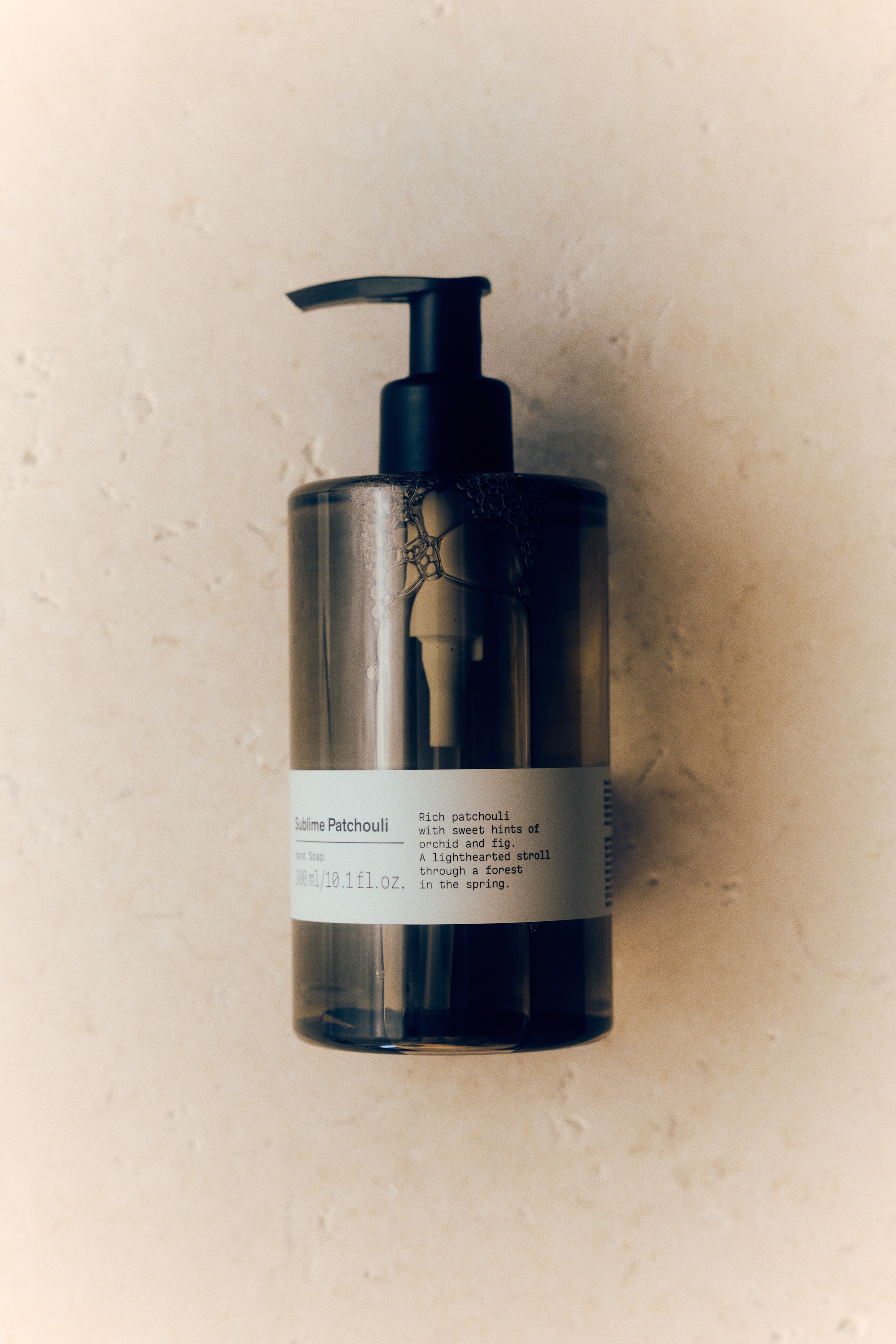 A front-facing view of a translucent, amber-tinted cylindrical hand soap bottle showing a black pump dispenser at the top and containing a clear, light-brown liquid. A white label wrapped around the lower portion displays black text, with the internal white pump mechanism visible through the container.