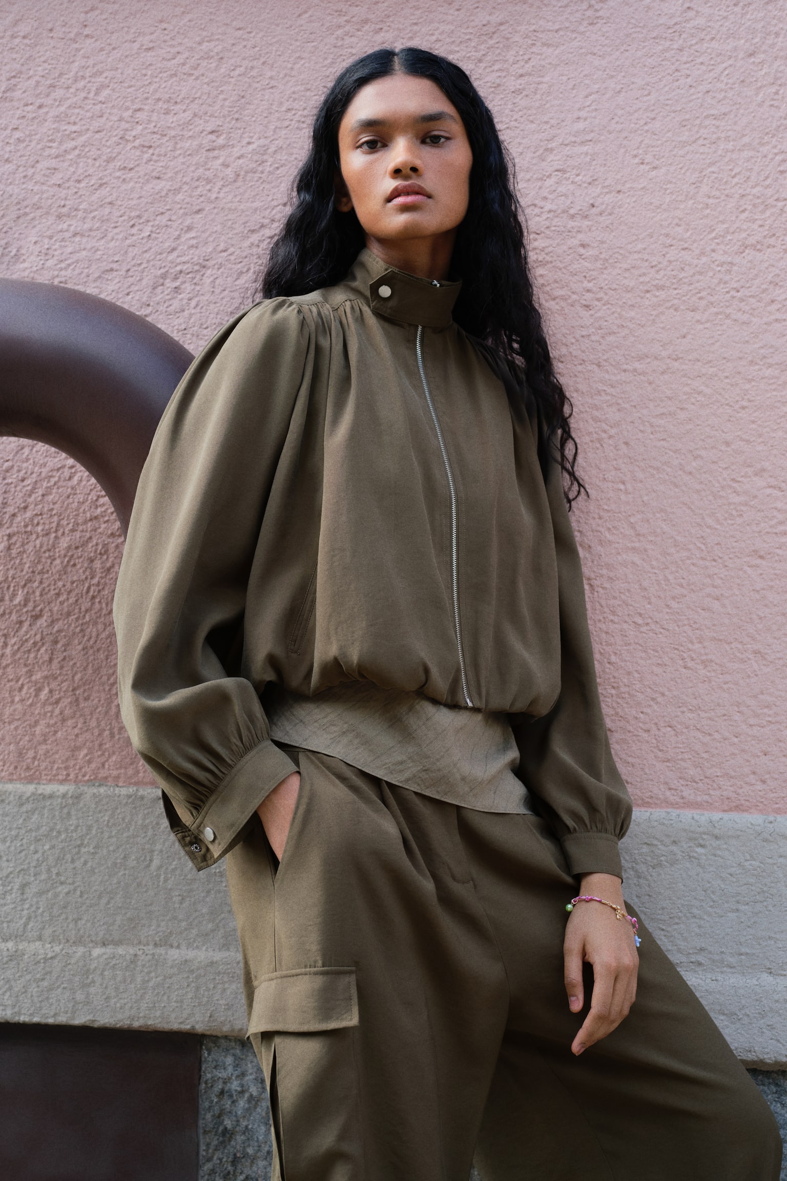 An olive green cropped jacket features a high stand-up collar fastened by a snap button and a central silver-toned zip. It displays gathered detailing at the shoulders and above the cuff bands, along with slanted welt pockets and an elasticated hem. - 1