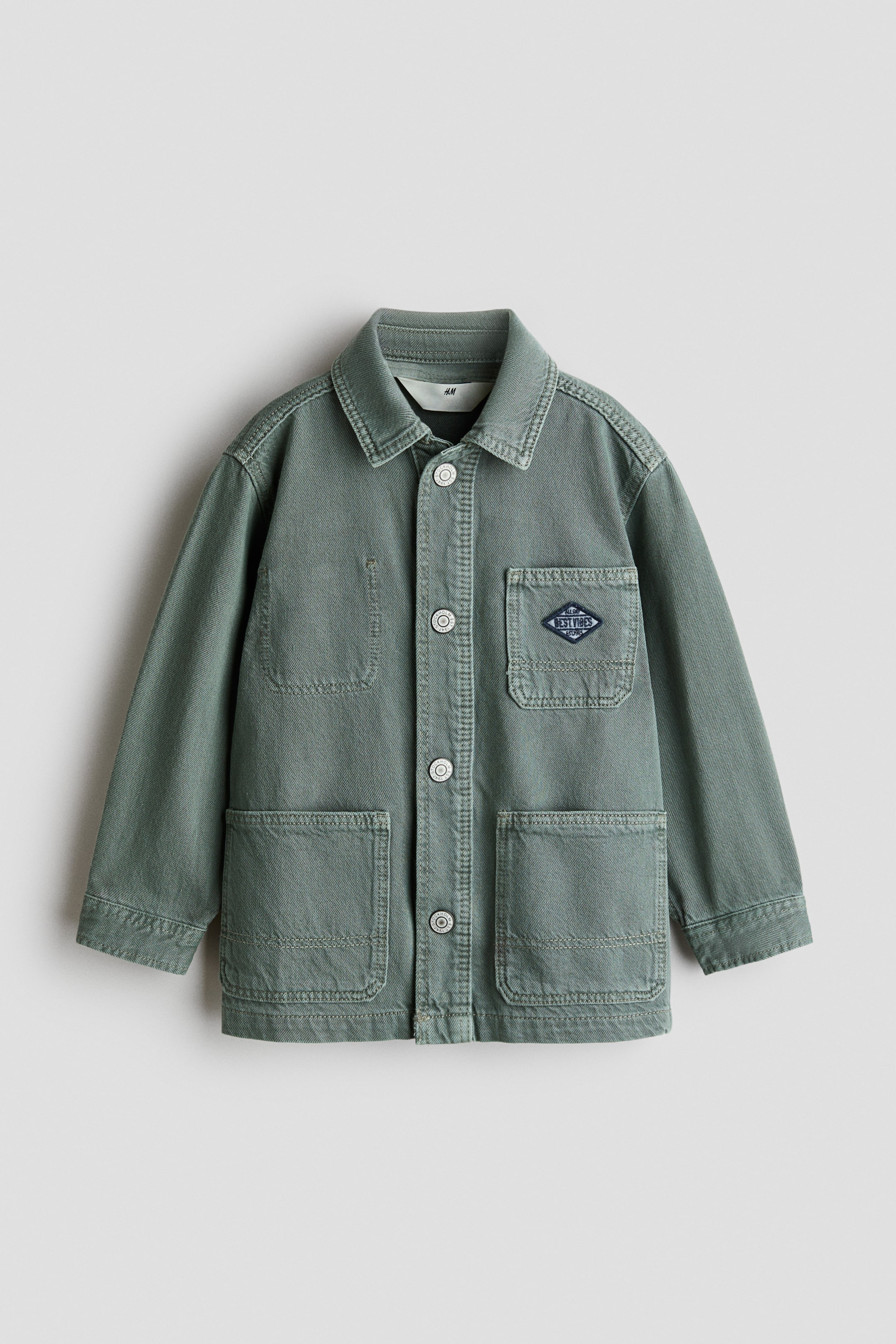 View larger image: A muted green utility jacket is facing front, featuring a pointed collar and a full button-front closure with five light-colored buttons. It is displaying four patch pockets on the front, two at the chest and two at the bottom, with a diamond-shaped patch reading "BEST VIBES" adorning the right chest pocket.