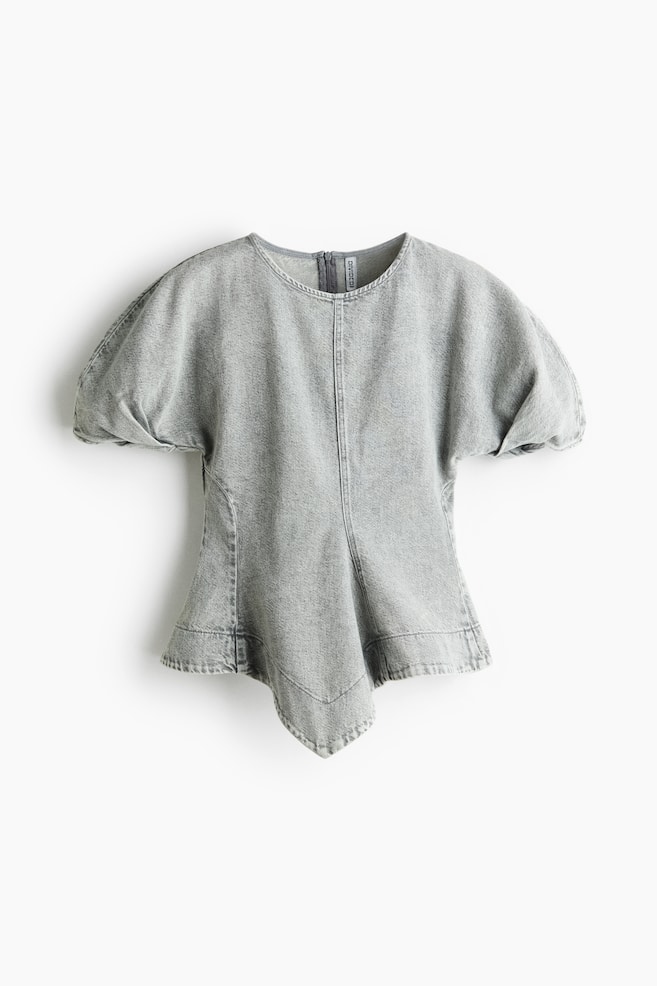 Eine hellgraue Denim-Bluse ist von vorne zu sehen und verfügt über einen runden Ausschnitt, kurze Puffärmel mit plissierten Manschetten und vertikale formgebende Nähte. Das Kleidungsstück ist an der Taille tailliert und läuft in eine Peplum mit spitzem, asymmetrischem Handkerchief-Saum aus. - 1