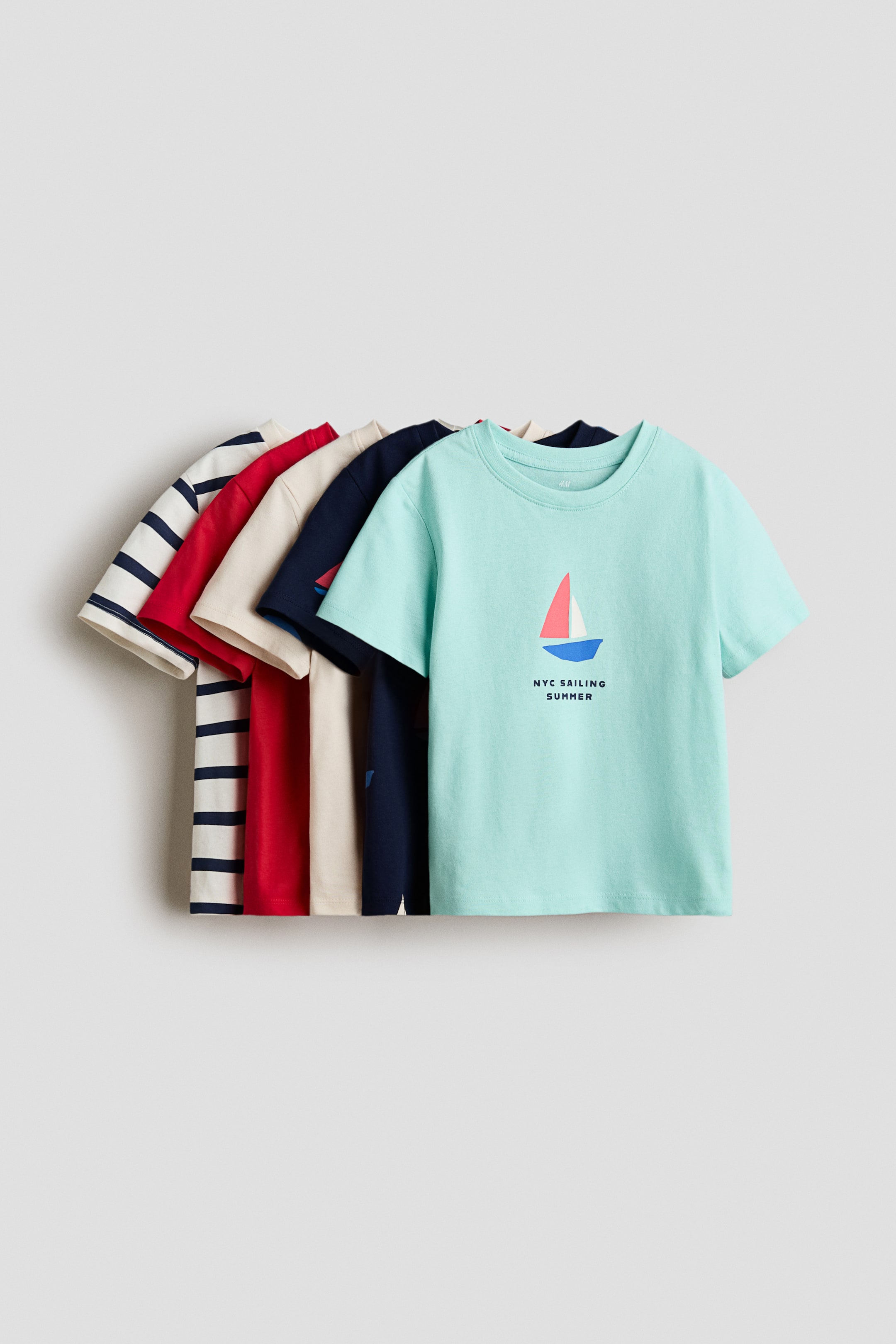 View larger image: A collection of classic t-shirts is on display, neatly stacked with their fronts facing forward. The foremost t-shirt is a light blue shade, featuring a round, rib-trimmed neckline and adorned with a sailboat print along with the text 'NYC SAILING SUMMER' on its front. Behind it, other t-shirts in hues of red, beige, dark blue, and navy stripes are layered, creating a visually appealing ensemble.