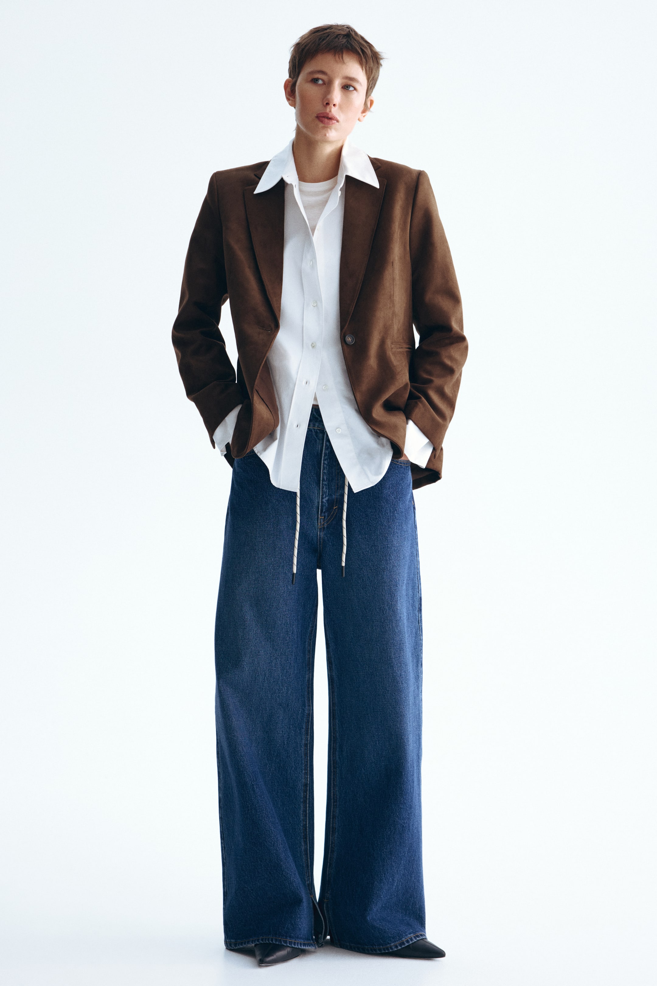 Ver imagem maior: Um blazer castanho com efeito de camurça está a ser usado, apresentando lapelas de entalhe e um fecho de botão único. Apresenta uma silhueta estruturada a partir de uma vista frontal ligeiramente angulada, usado aberto sobre uma camisa branca com colarinho.