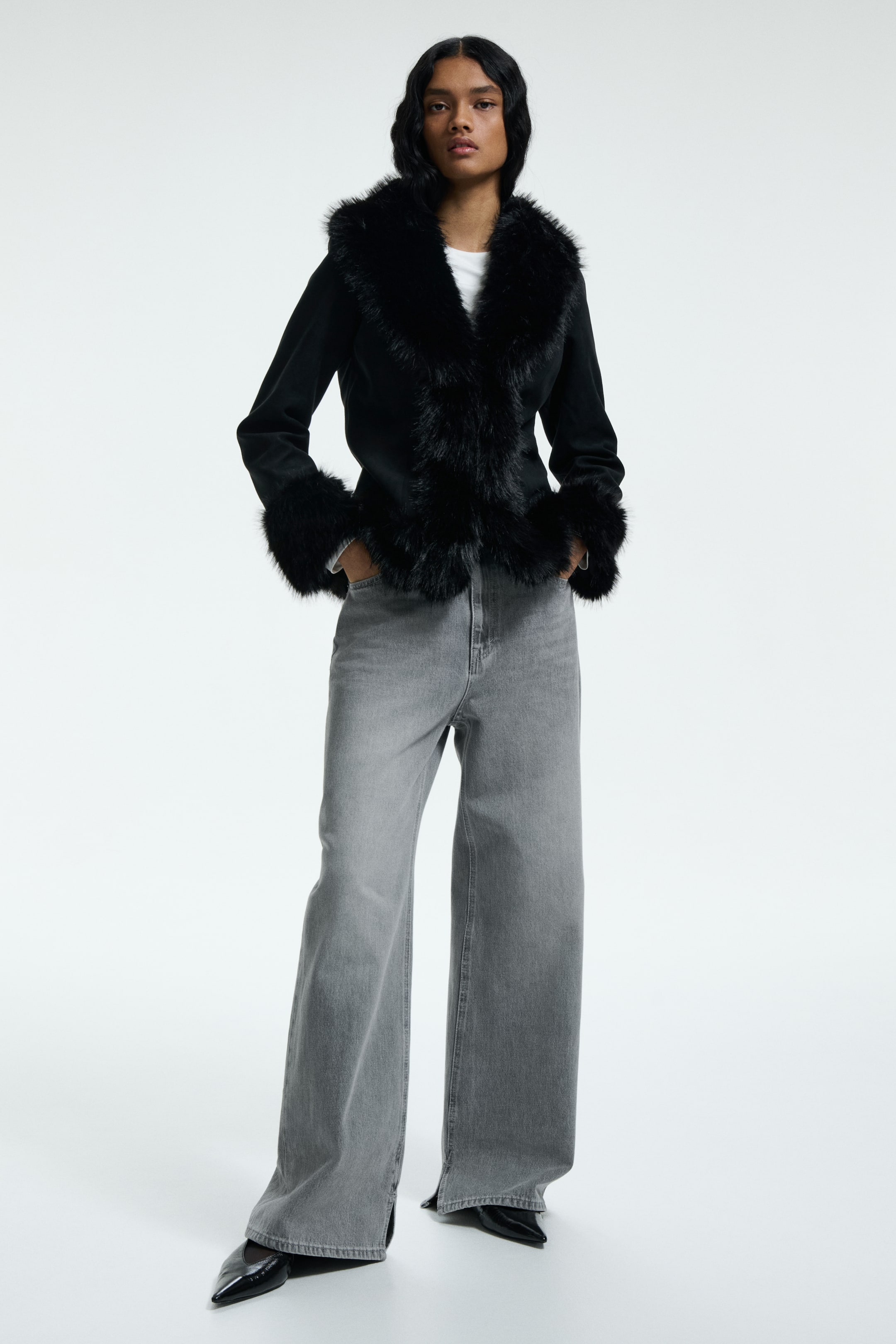 Agrandir l'image: Une veste noire est présentée de face, avec un col en fausse fourrure noire et une garniture qui s'étend jusqu'à l'ouverture avant et le long des poignets. Elle est portée ouverte sur un haut blanc, mettant en valeur sa longueur raccourcie.