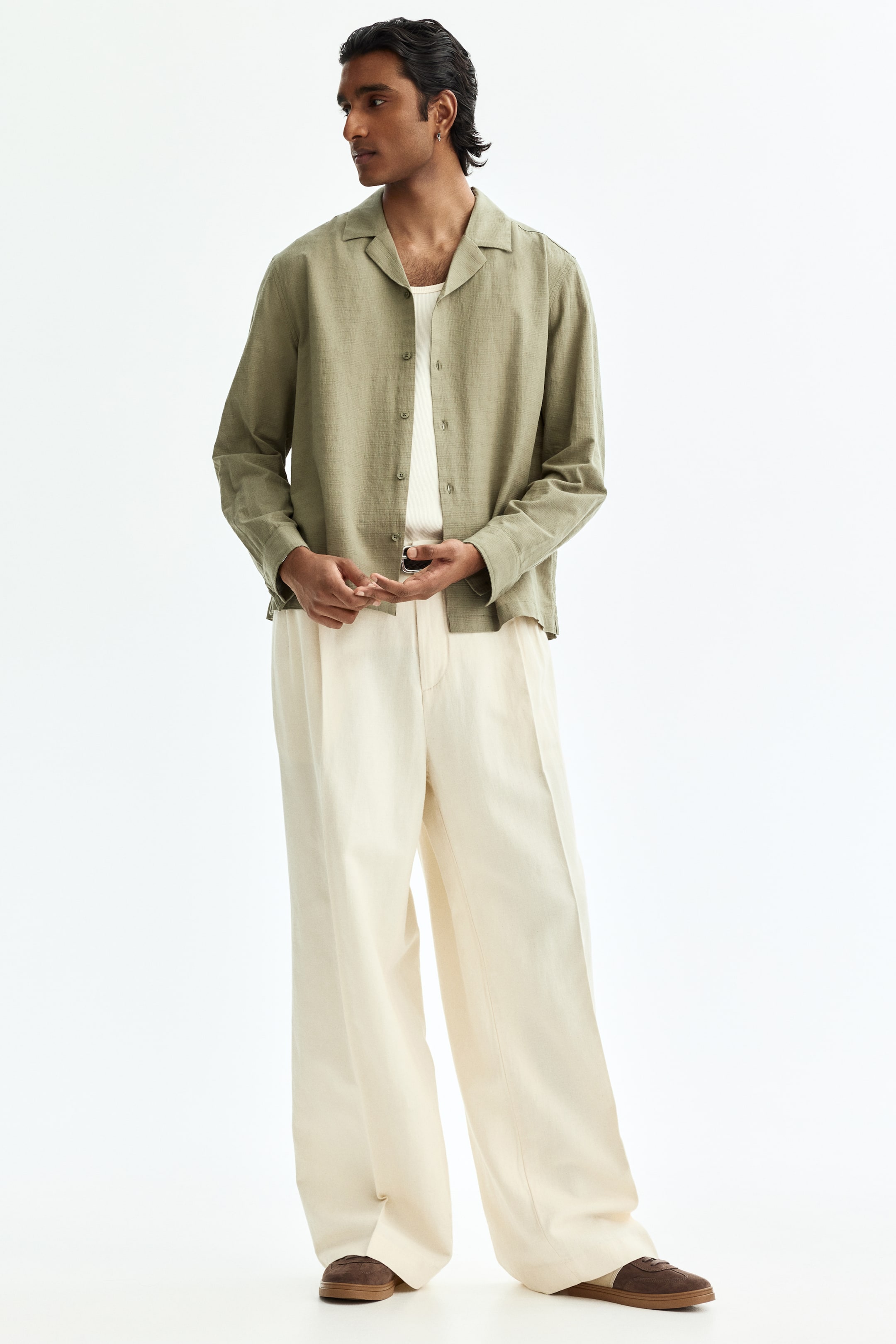 View larger image: An olive green long-sleeved shirt with a camp collar is worn open over a white undershirt, its relaxed fit complementing cream wide-leg pleated trousers. The trousers fall loosely over brown and cream shoes.