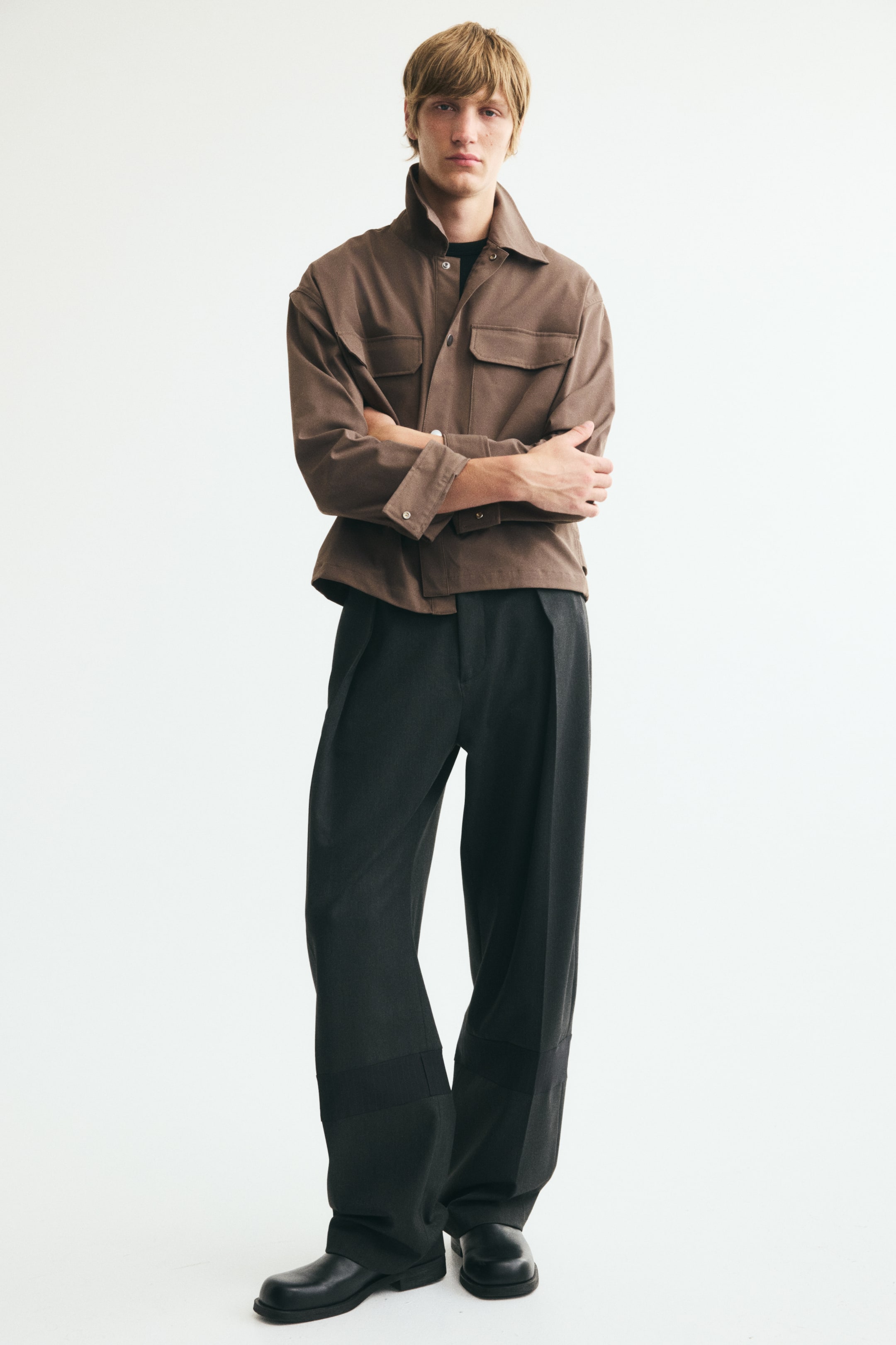 Voir une image plus grande: Une veste utilitaire structurée brun chocolat, dotée d’un col pointu, de poches poitrine à rabat et de poignets boutonnés, est portée par le modèle. La veste est superposée à un haut noir à col rond et agencée à un pantalon plissé à jambe large de couleur gris foncé.