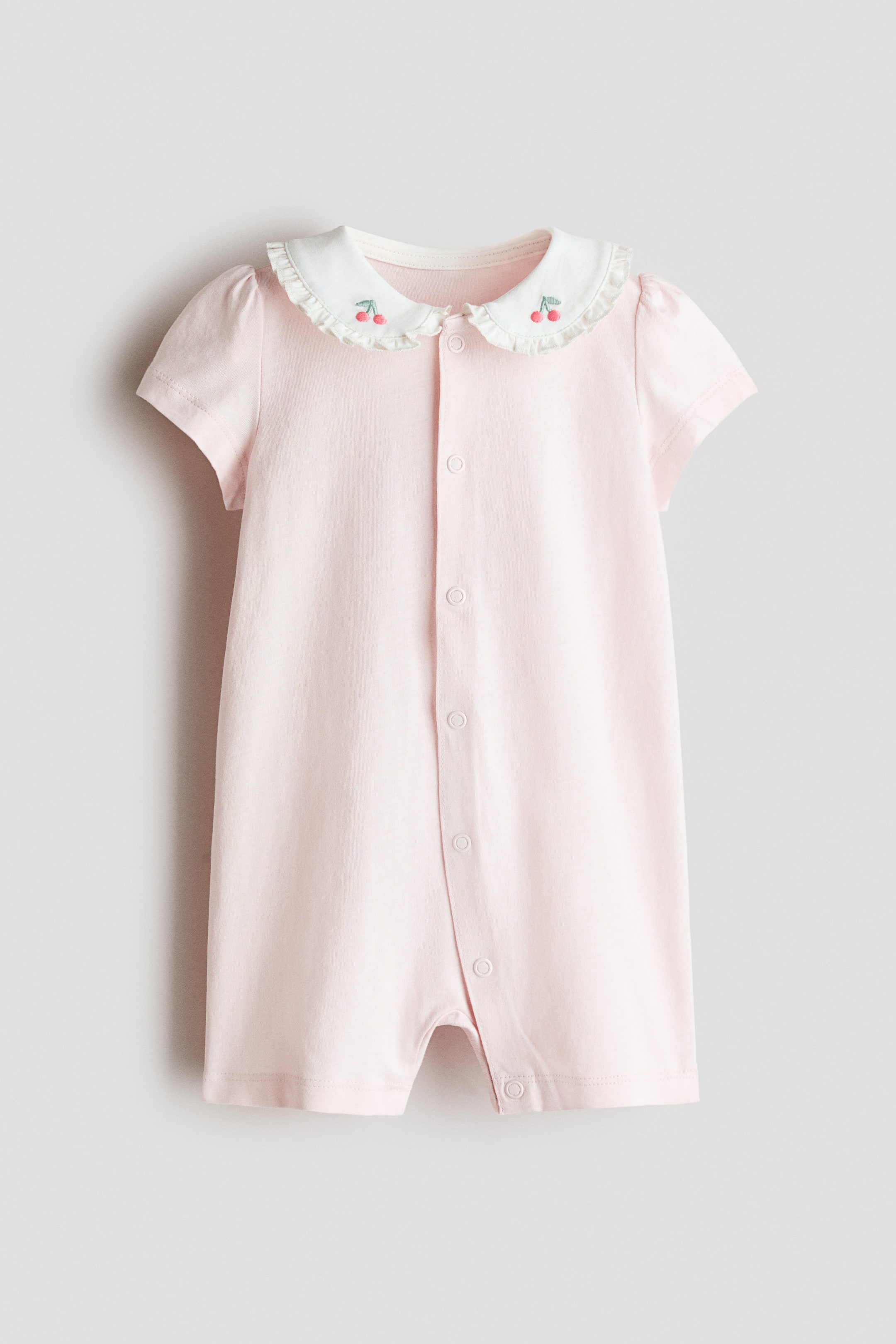 View larger image: A light pink baby romper featuring a white ruffled Peter Pan collar with cherry embroidery on the collar points. It also presents short sleeves and a full-length snap button placket down the front.