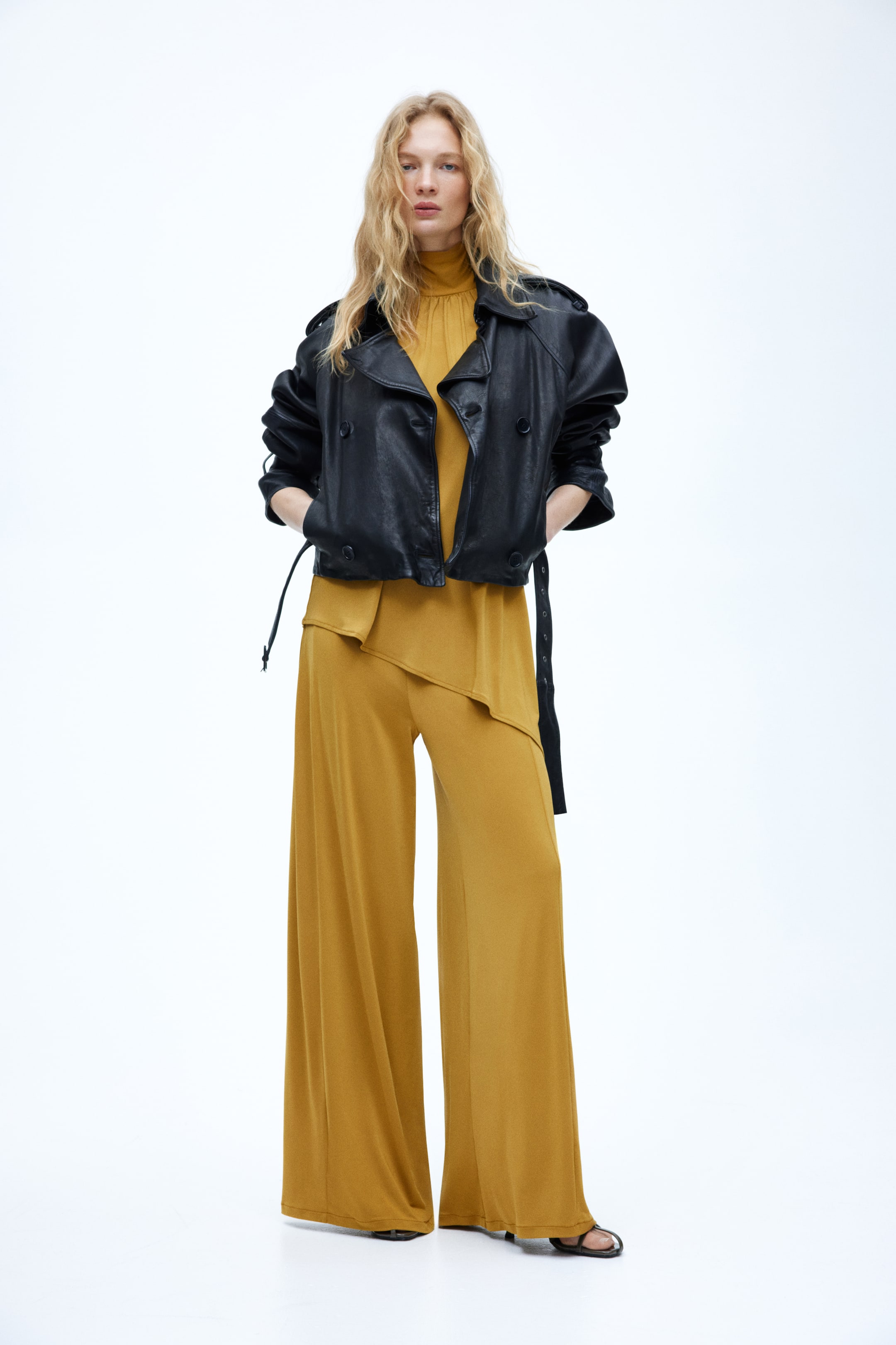 View larger image: A cropped black leather double-breasted jacket is worn, featuring wide lapels, shoulder epaulettes, and a belted hem with the belt hanging loosely. It is layered over a mustard-coloured high-neck top and wide-leg trousers.
