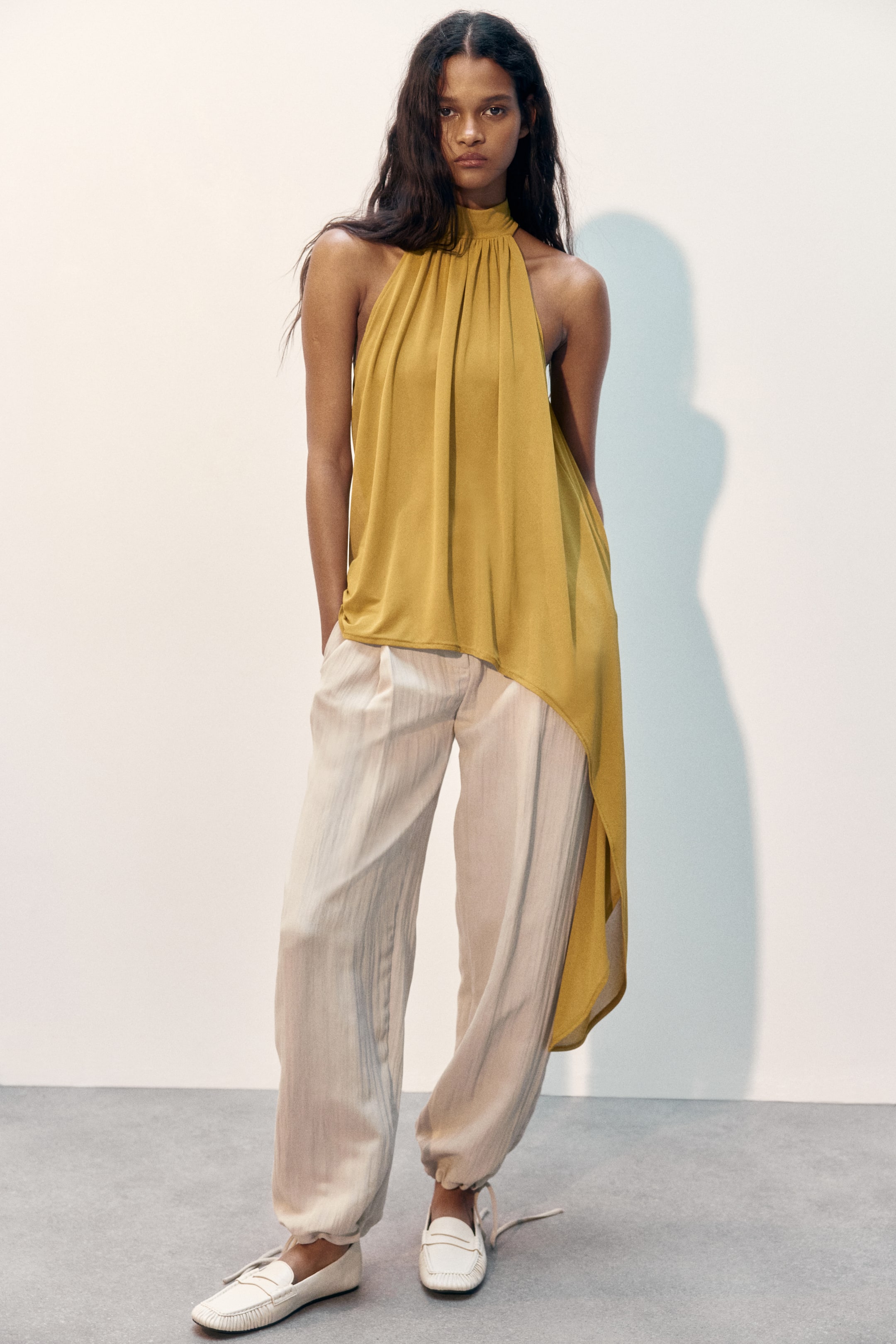 Ampliar la imagen: La modelo lleva puesto un top halter de ajuste holgado en color amarillo dorado. Presenta un cuello alto, frunces en la parte superior que crean un efecto suavemente drapeado y un dobladillo asimétrico.