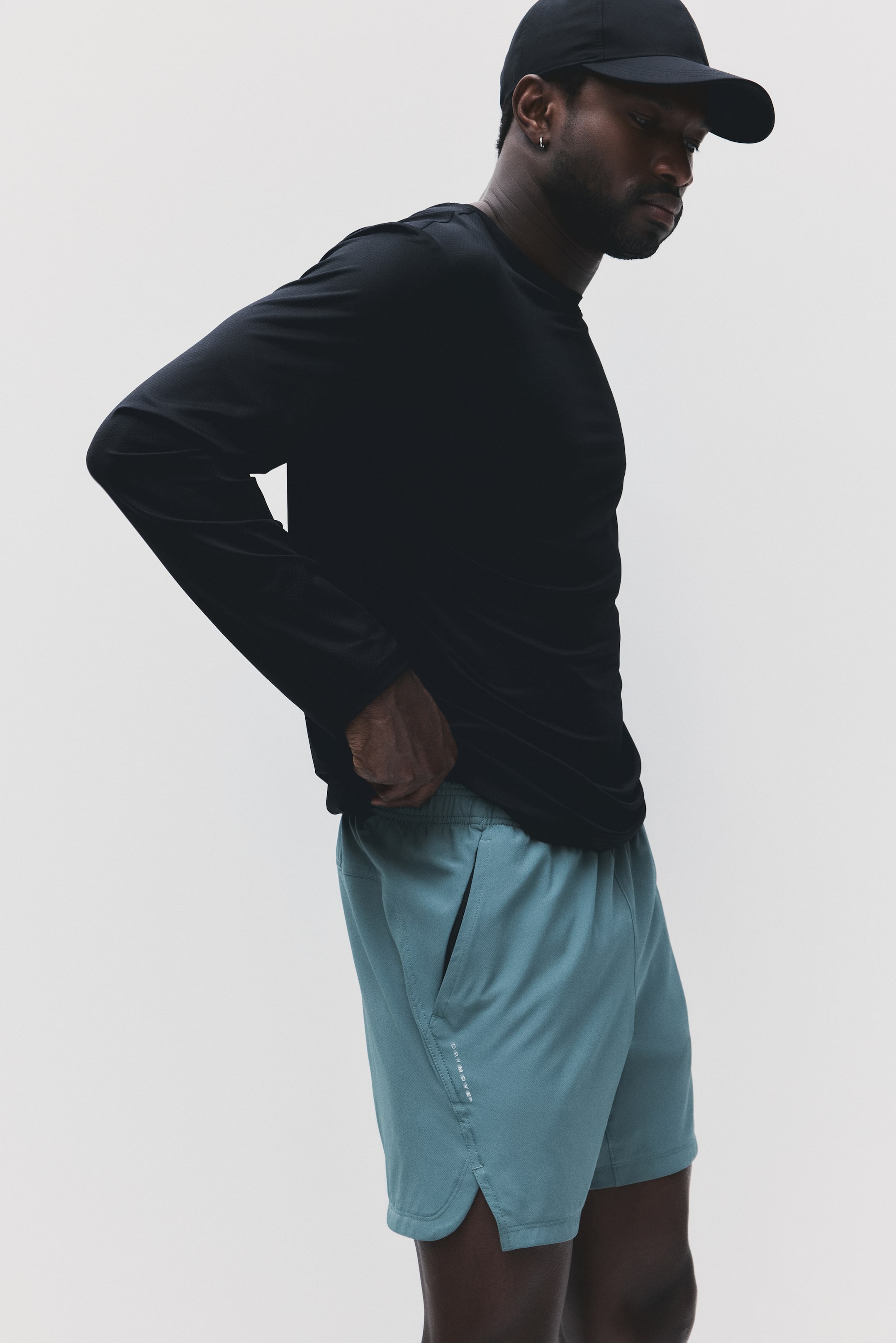 Voir une image plus grande: Un chandail noir à manches longues et à col rond est porté, son ourlet partiellement rentré dans un short de sport turquoise, les deux visibles sous un léger angle latéral. Le short présente une taille élastique, des poches latérales, un ourlet fendu et un petit logo vertical blanc sur la cuisse gauche.