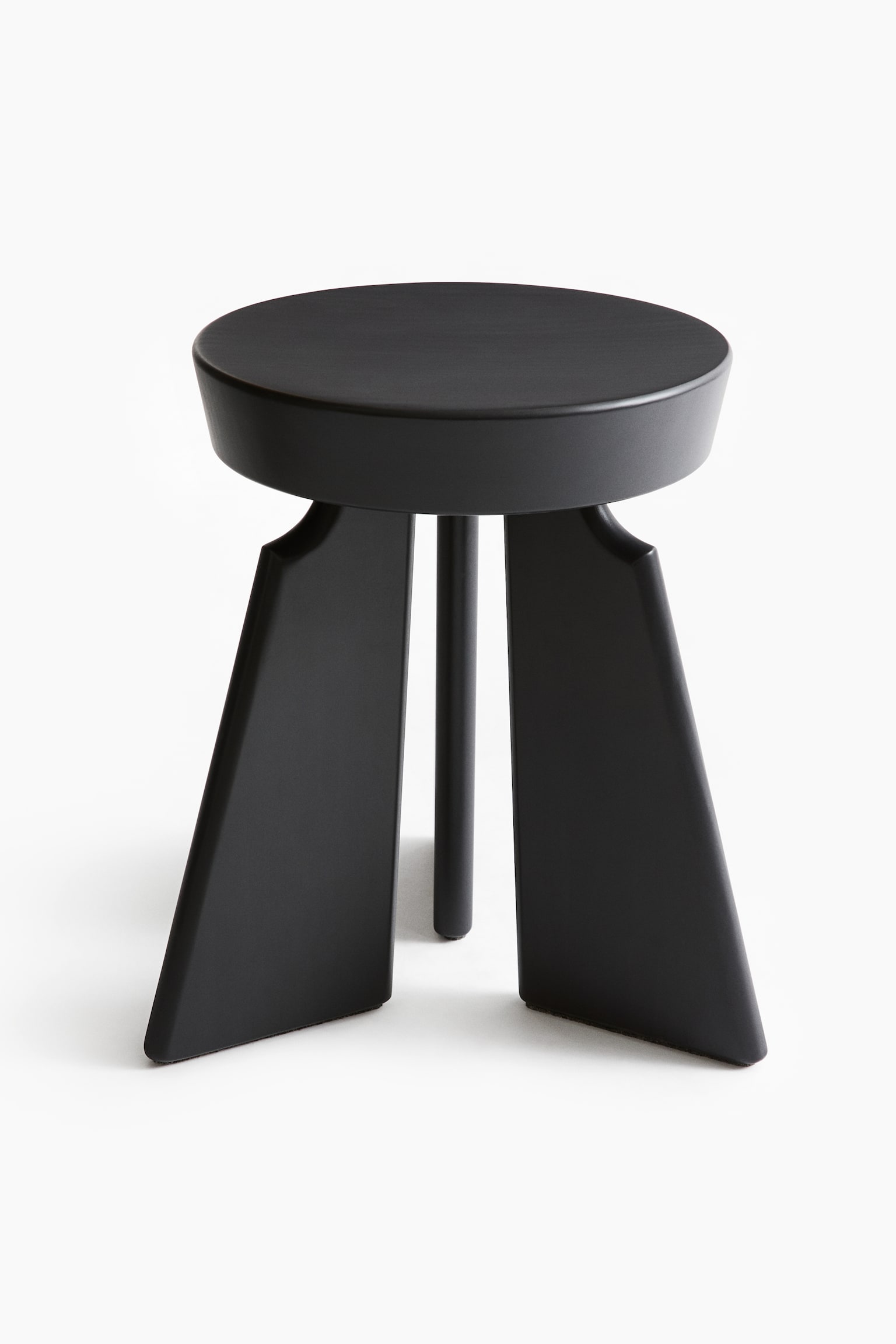 A black stool is shown with a round, flat seat and a subtle bevelled edge. It is supported by three legs, with two broad, trapezoidal legs angling outwards from the seat and one thinner, cylindrical leg visible behind them. - 1
