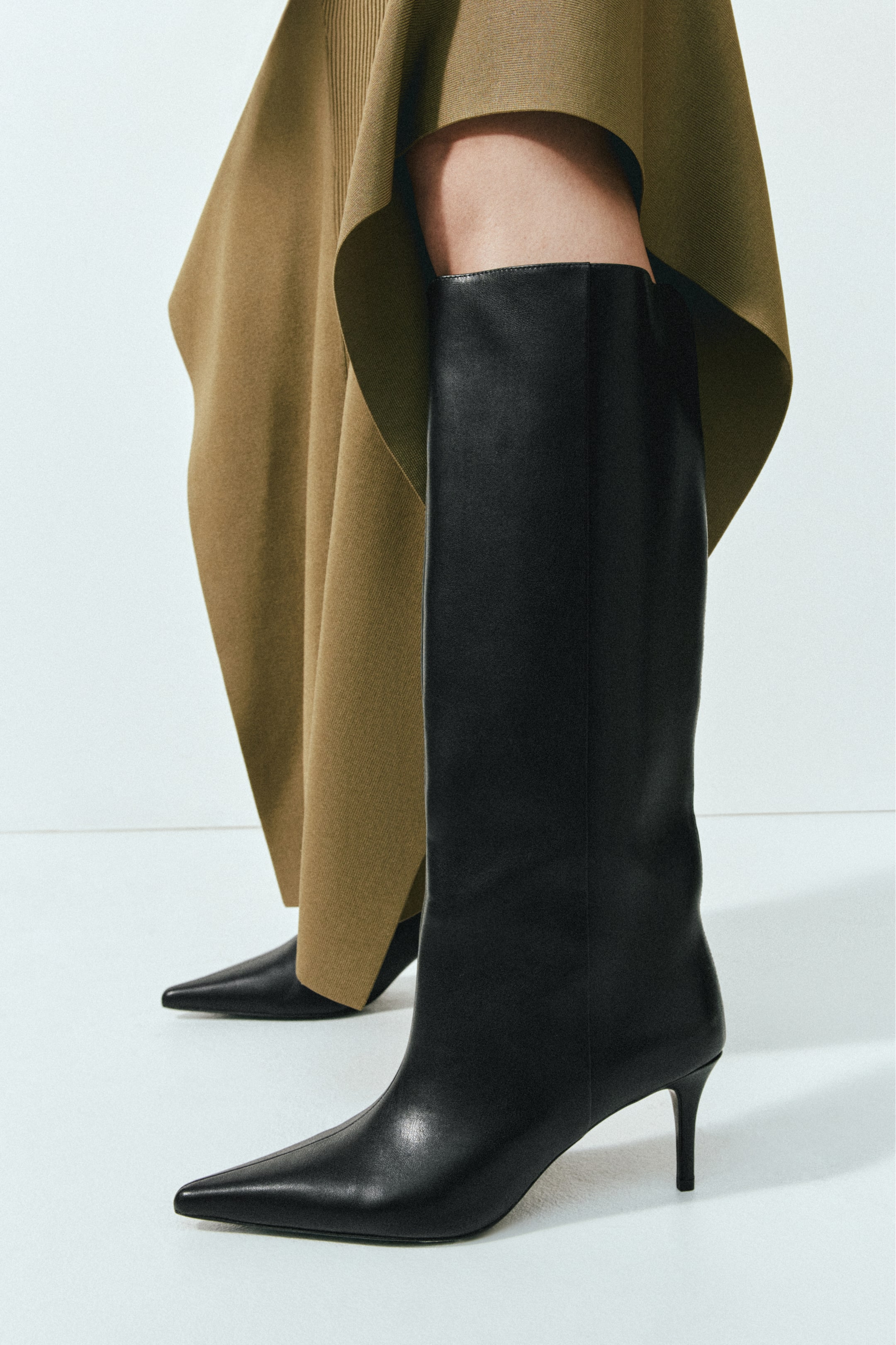 View larger image: Black knee-high boots featuring pointed toes, narrow heels and wide shafts are being worn by the model. The boots are paired with a draped, olive-green knitted skirt.