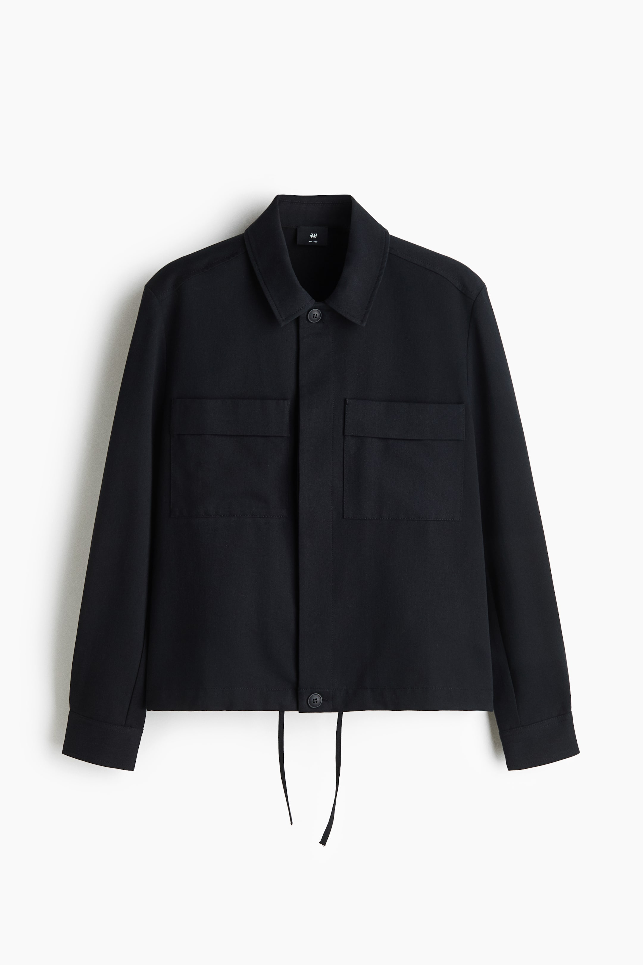 Größeres Bild anzeigen: Eine schwarze Jacke mit langen Ärmeln ist in Vorderansicht zu sehen und verfügt über einen klassischen Spitzkragen, eine verdeckte Knopfleiste und zwei große Brusttaschen mit Patten. Das Kleidungsstück ist mit einem geraden Saum gestaltet, von dessen Mitte zwei dünne schwarze Kordeln herabhängen.