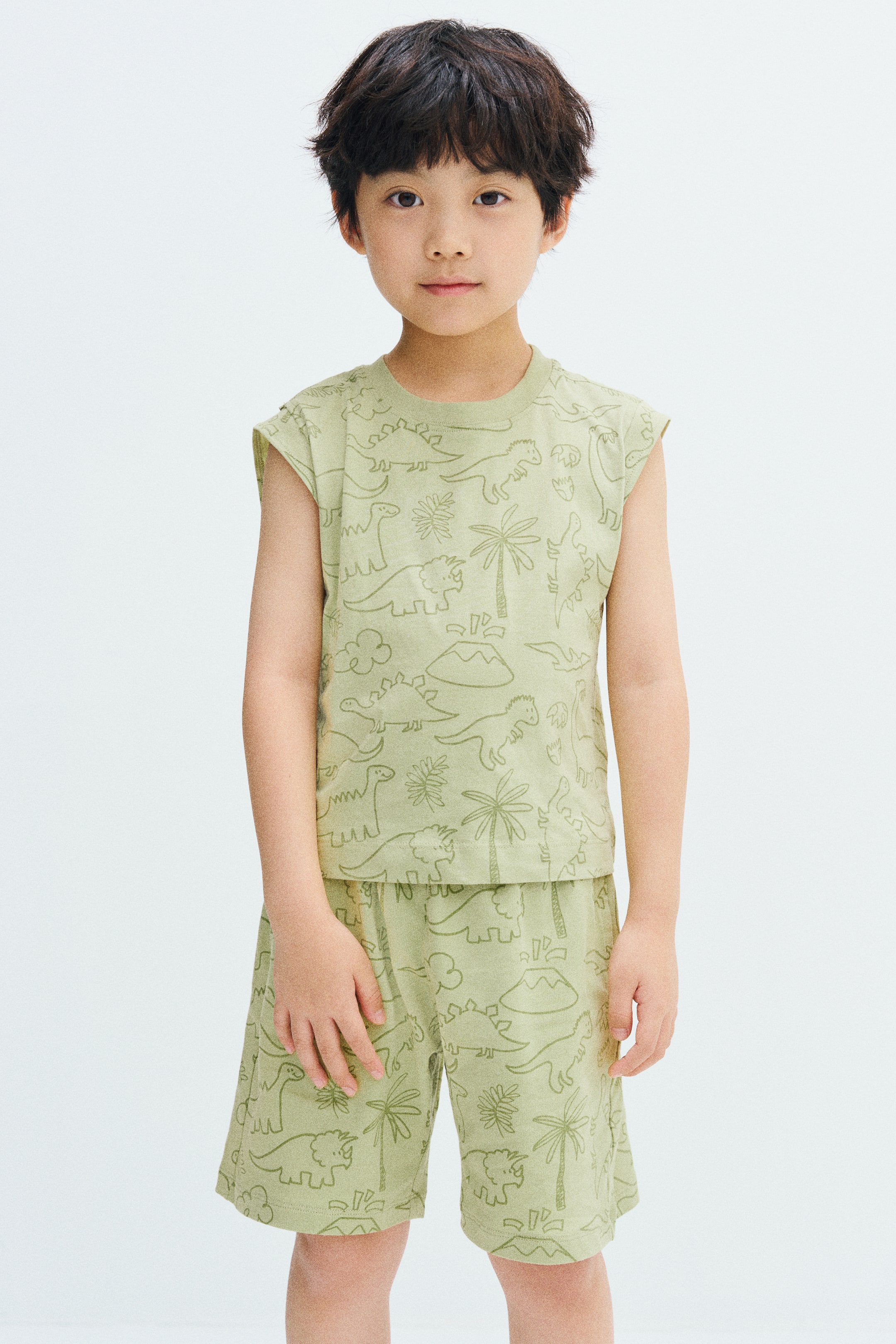 View larger image: The model is wearing knee-length shorts in light green with a printed dinosaur and palm tree pattern. The shorts are styled with a matching sleeveless T-shirt featuring the same design.