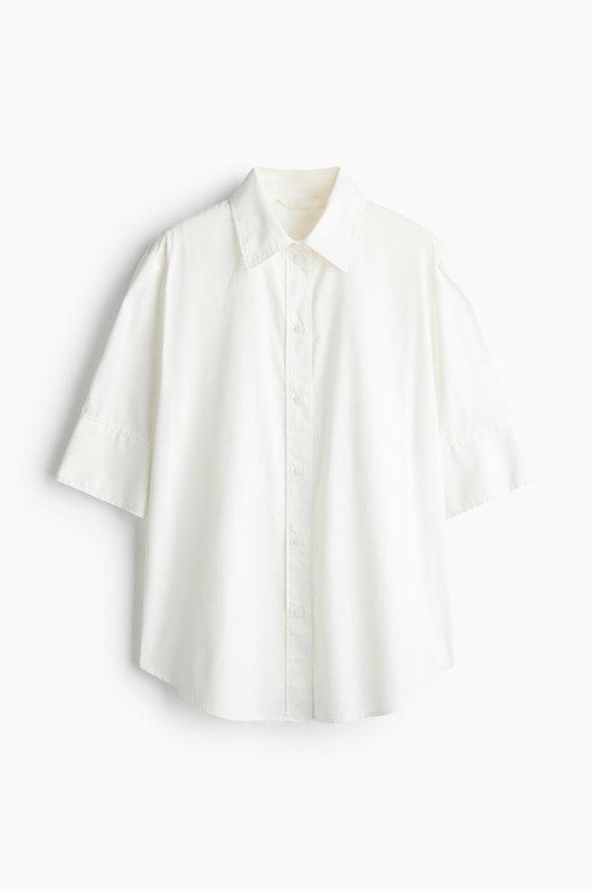 A white cotton poplin shirt is shown facing front, featuring a classic pointed collar, a button-down front, and wide, elbow-length sleeves with subtle pleat details. The shirt is hanging flat, displaying a relaxed silhouette and a rounded hem. - 1