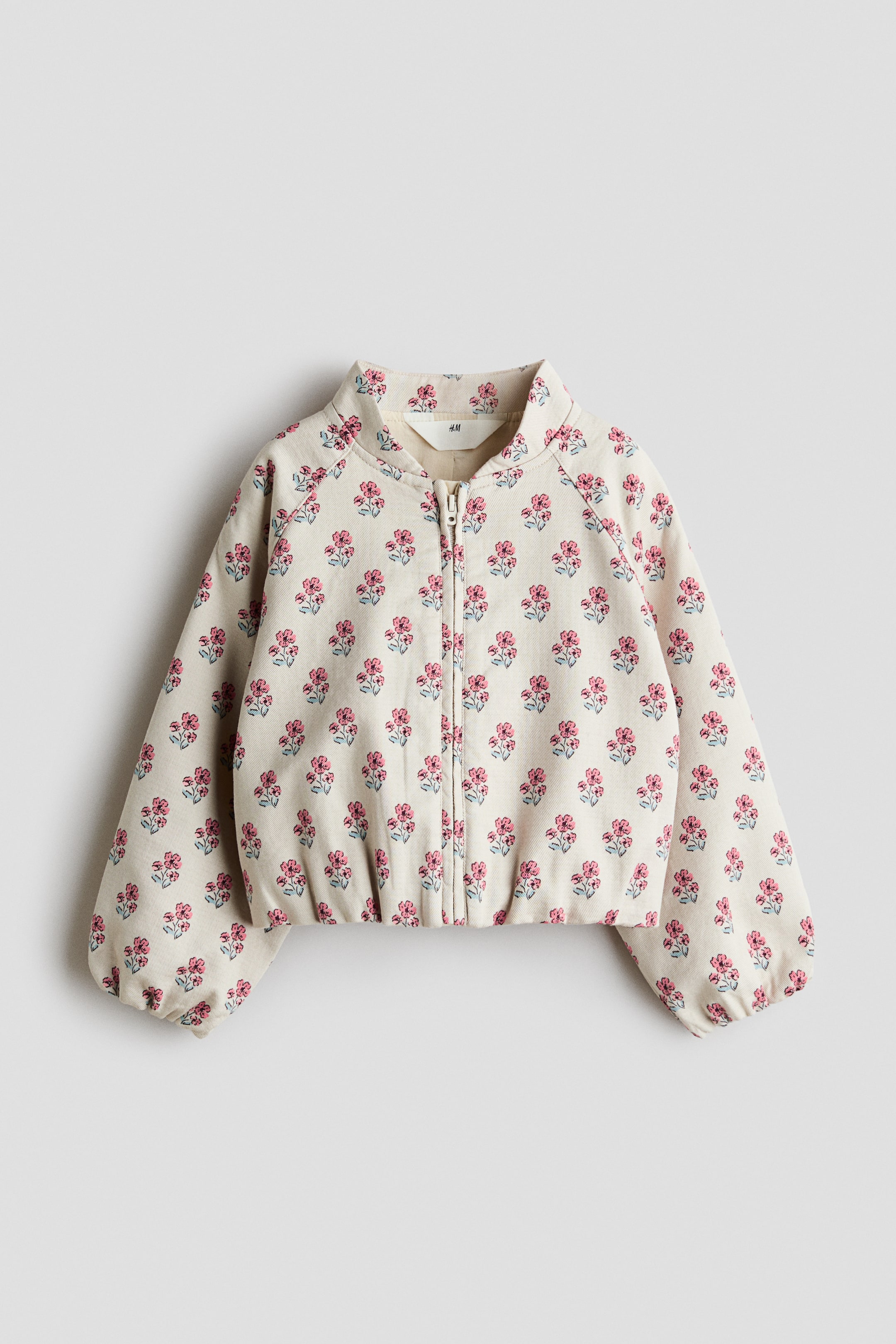 View larger image: A beige jacket features an all-over pattern of pink flowers with green leaves, a full-length front zip, and a stand-up collar, along with long raglan sleeves with gathered cuffs and an elasticated hem.