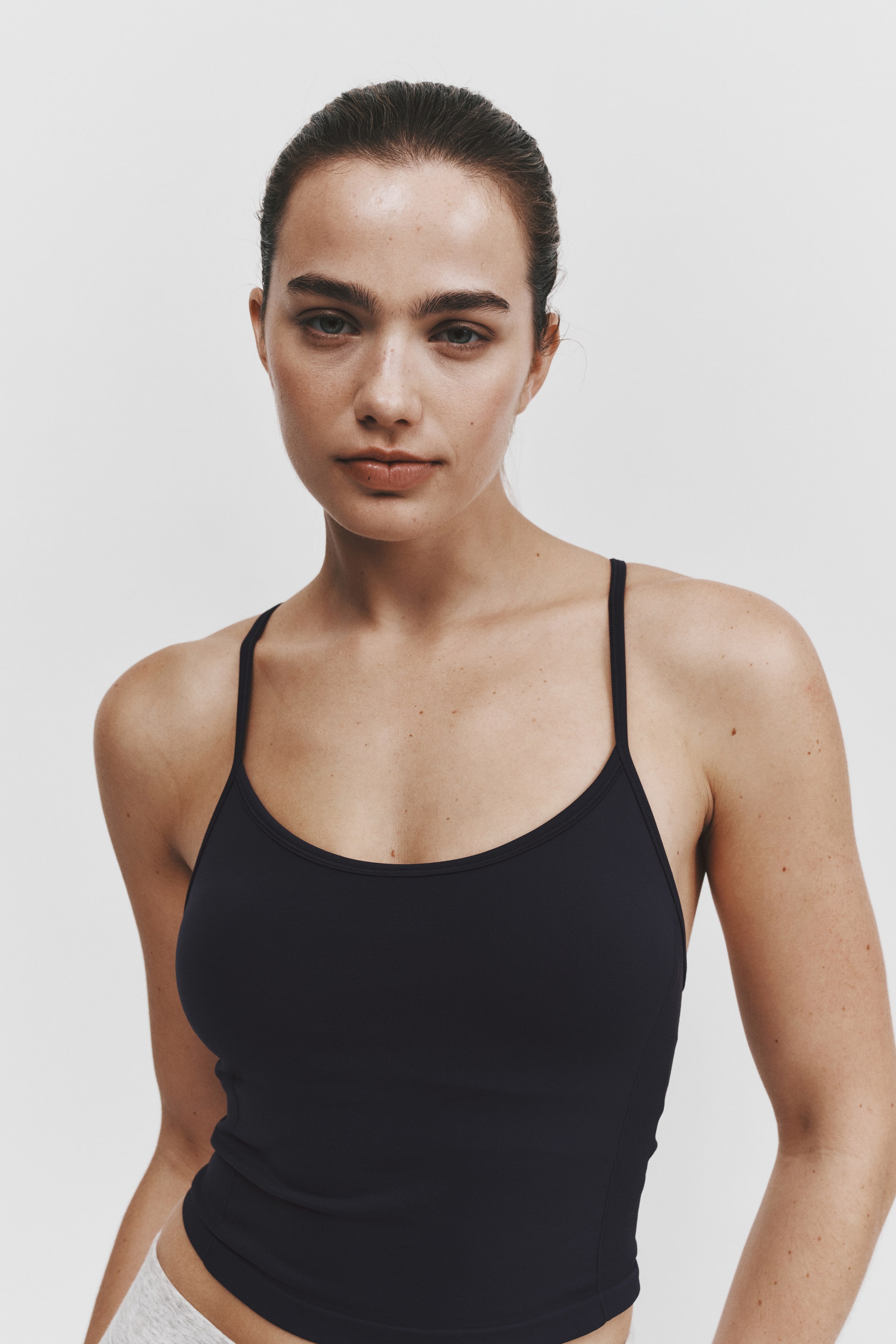 Agrandir l'image: Un haut de sport noir, ajusté, est porté de face, avec de fines bretelles spaghetti et un col rond échancré. Ce haut sans manches épouse le buste et s’arrête à la taille, juste au-dessus d’un bas gris chiné clair.