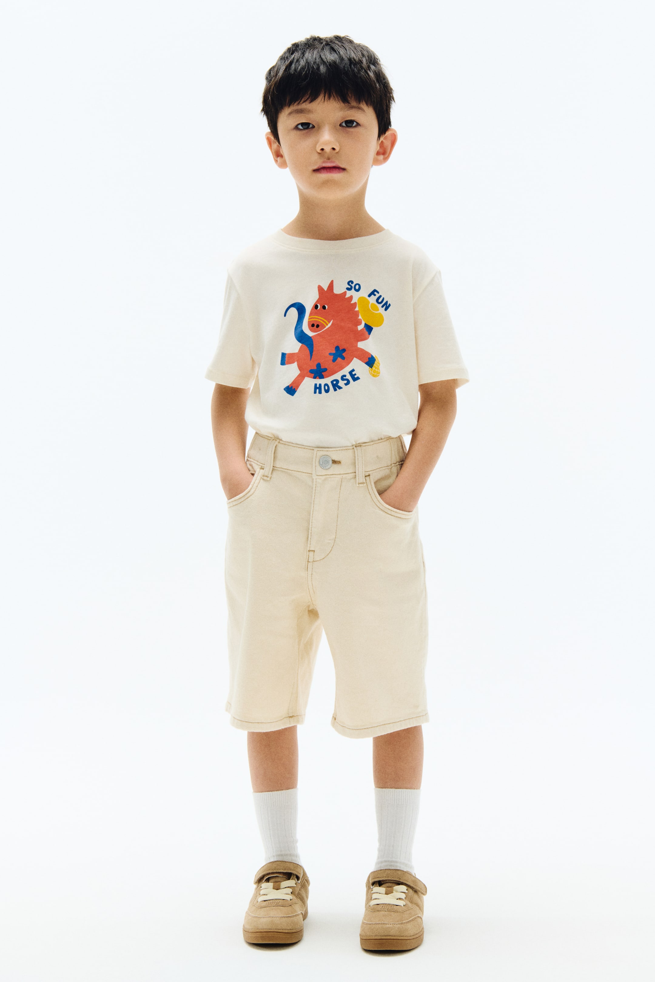 View larger image: An off-white short-sleeved T-shirt featuring a colourful graphic of a red horse with blue text reading “so fun horse”. It is paired with light beige shorts with front pockets, white crew socks, and tan low-top sneakers with Velcro straps.