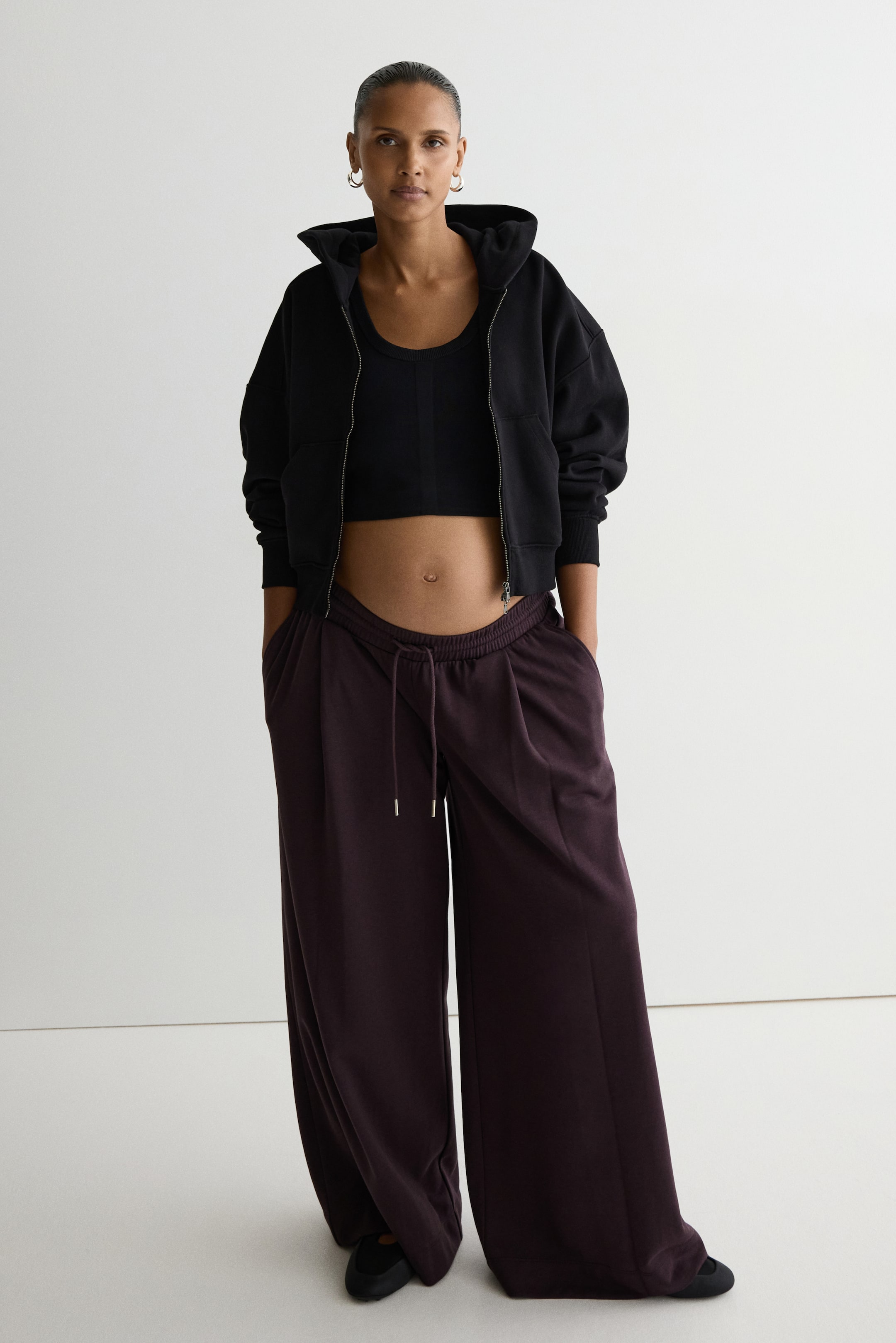 View larger image: Dark plum-colored loose-fit sweatpants are worn by the model, who places her hands in the diagonal side pockets. The pants feature a low waist with smocked elastic, a drawstring, and wide legs with an inverted box pleat at the front, styled with a black crop top and an open black zip-up hoodie.
