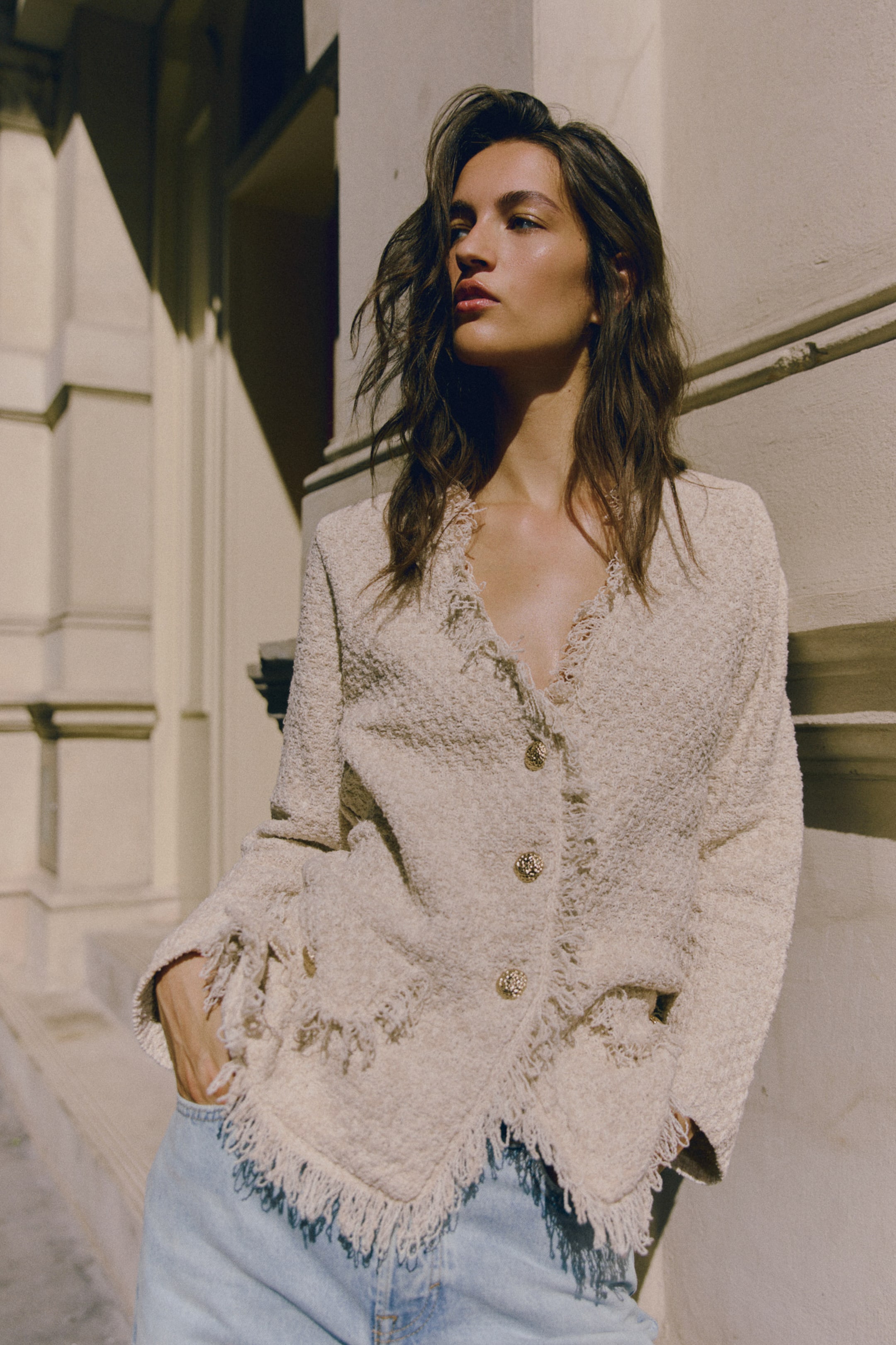 Agrandir l'image: Un long cardigan de couleur crème en maille douce à tressage panier est présenté de face, avec un col en V, des manches longues et des boutons en métal texturé sur le devant. Le cardigan met en valeur des bords fortement effilochés le long de l’encolure, des rabats de poches décoratifs et l’ourlet.