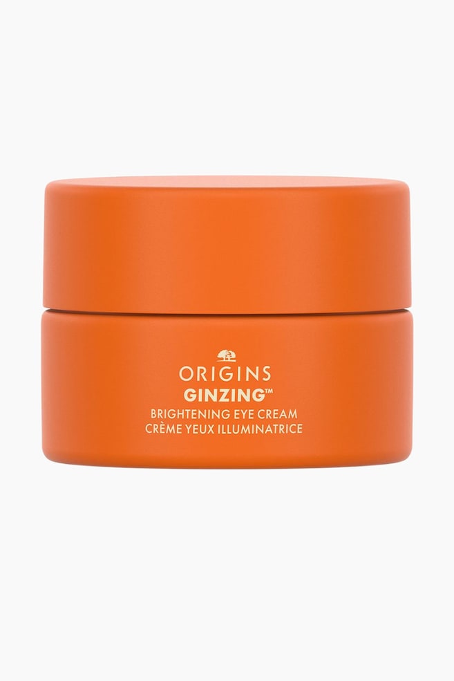 En lille, cylindrisk mat orange krukke med Origins GinZing Brightening Eye Cream stÃ¥r oprejst og vender fremad med det matchende orange lÃ¥g lukket. Forsiden af krukken viser cremefarvet tekst, der lyder "ORIGINS GINZING? BRIGHTENING EYE CREAM" og "CRï¿½ME YEUX ILLUMINATRICE" under brandets trÃ¦l logo. - 1
