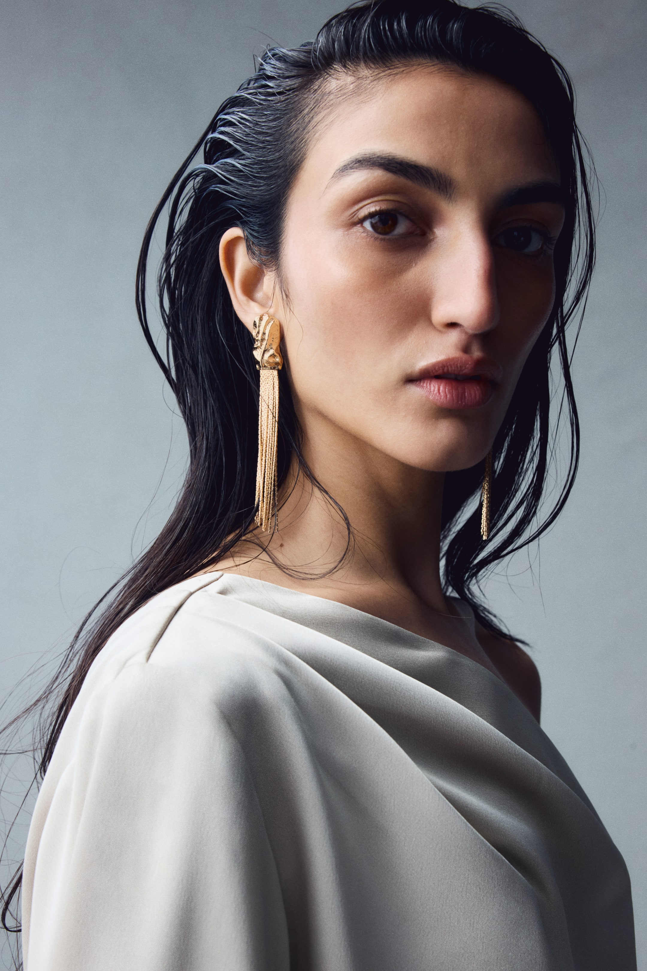 View larger image: Gold-toned metal dangle earrings featuring a textured, organic-shaped top piece and long, fine metal strands are worn by the model. The cascading strands fall toward the collarbone and are styled with a draped cream garment.