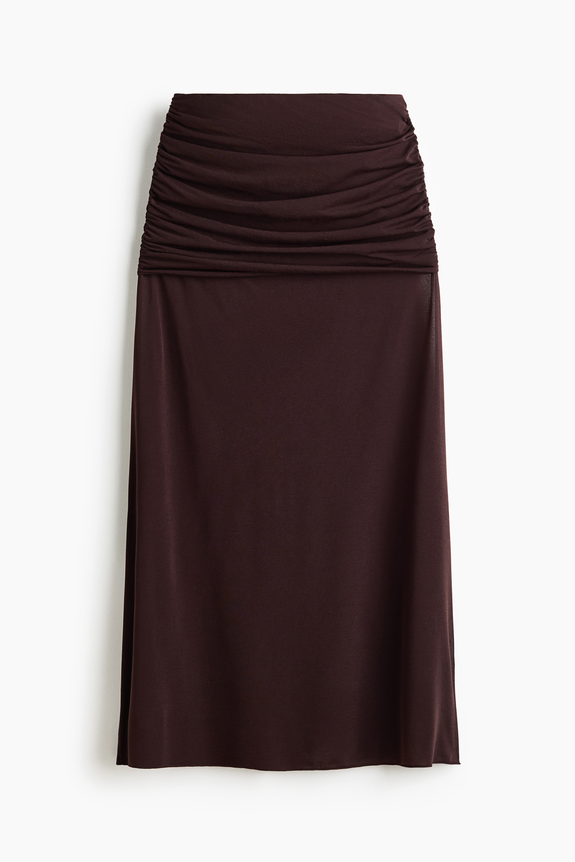A white jersey maxi skirt featuring a wide, gathered layer at the hips that creates a draped effect is worn by the model. The model is holding the side of the semi-sheer, flared hem of the skirt, styled with a matching sleeveless top.