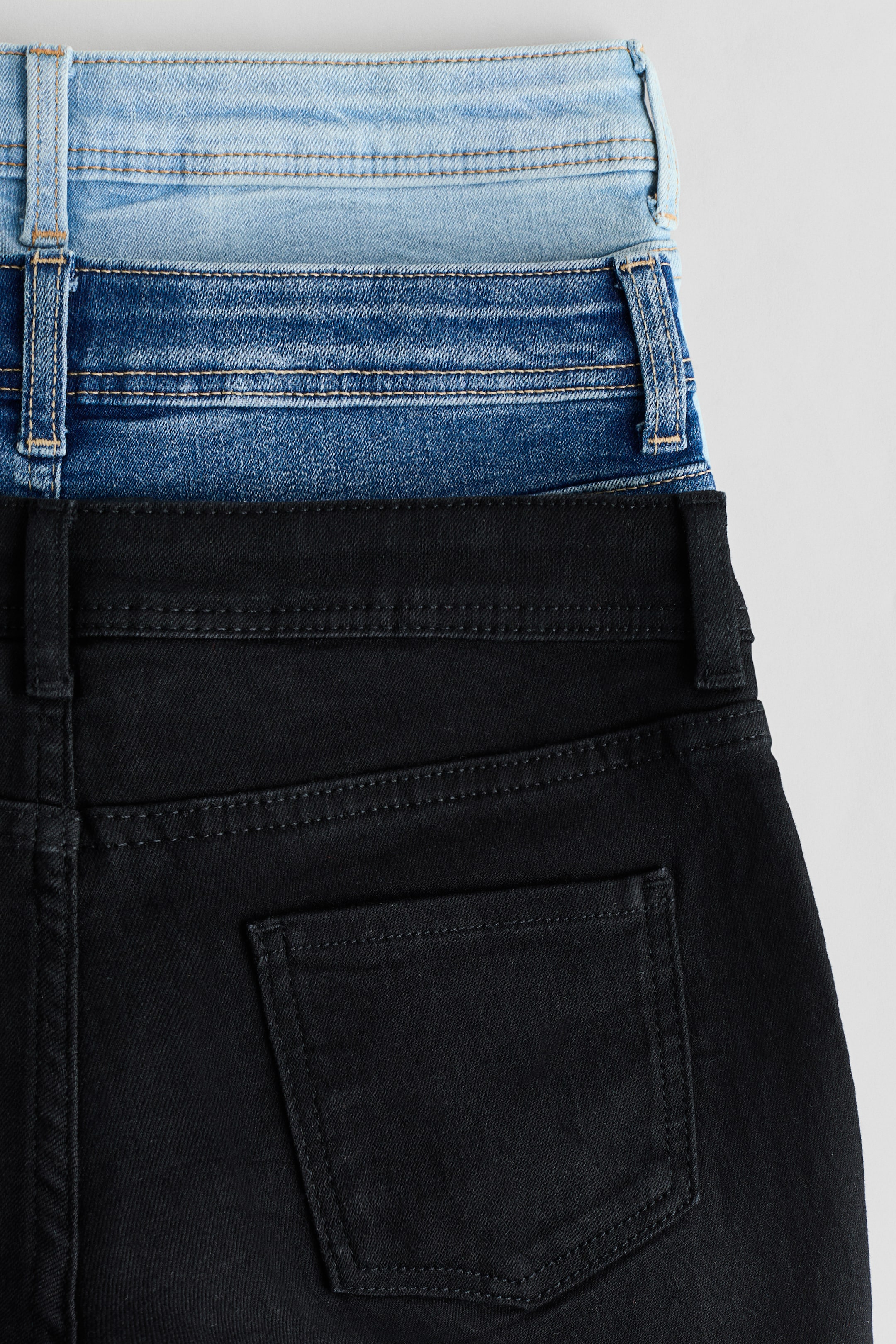 Ver imagen más grande: Los pantalones cortos de mezclilla de corte ajustado están mostrando su parte trasera, con tres pares apilados y visibles en azul claro, azul medio y negro. Cada par cuenta con una pretina con trabillas para cinturón, y los pantalones cortos negros muestran un bolsillo trasero abierto.