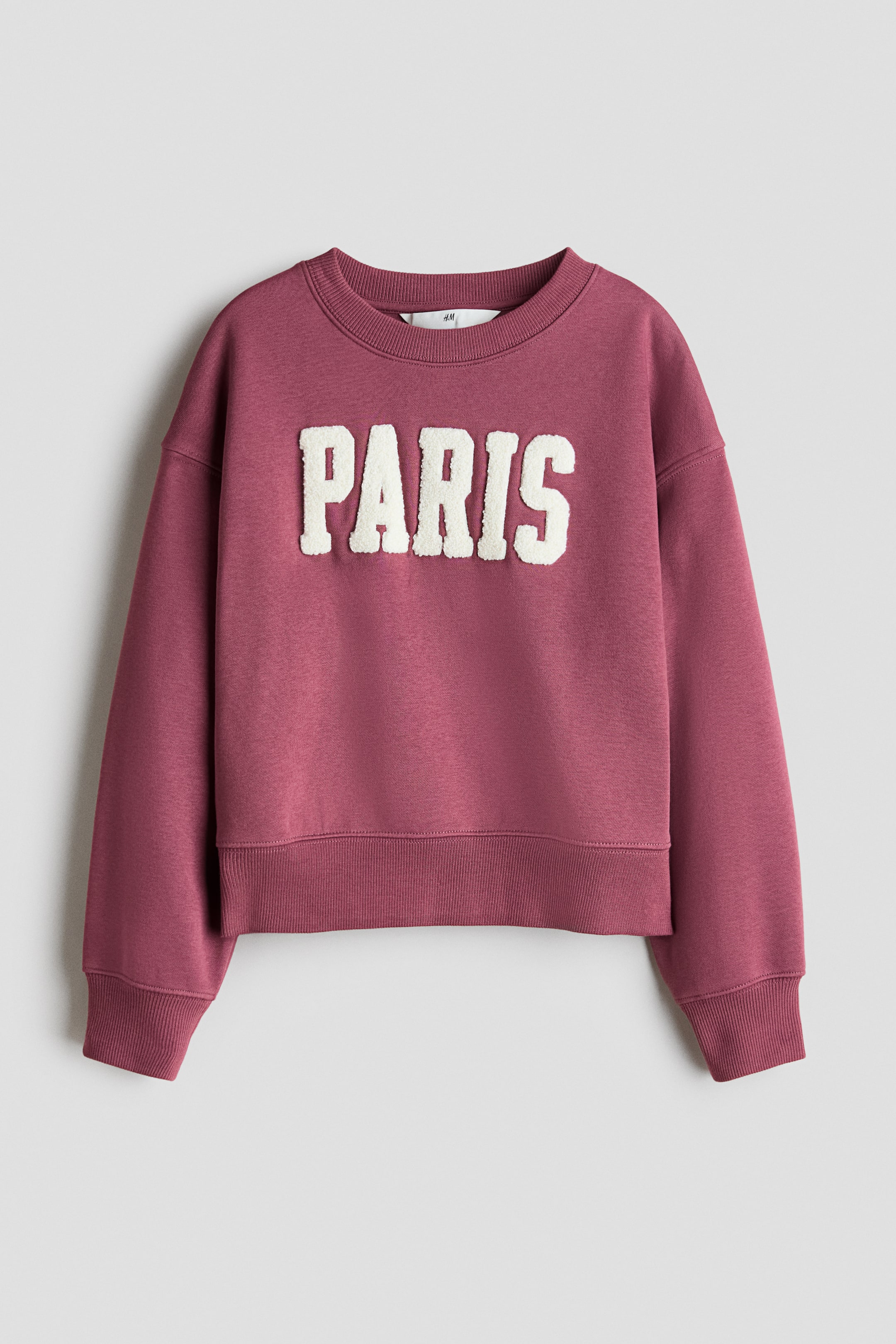 View larger image: A deep pink sweatshirt is showcased from the front, featuring a ribbed crew neck, long sleeves with ribbed cuffs, and a ribbed hem. The word "PARIS" appears in large, raised, off-white textured letters across the chest.