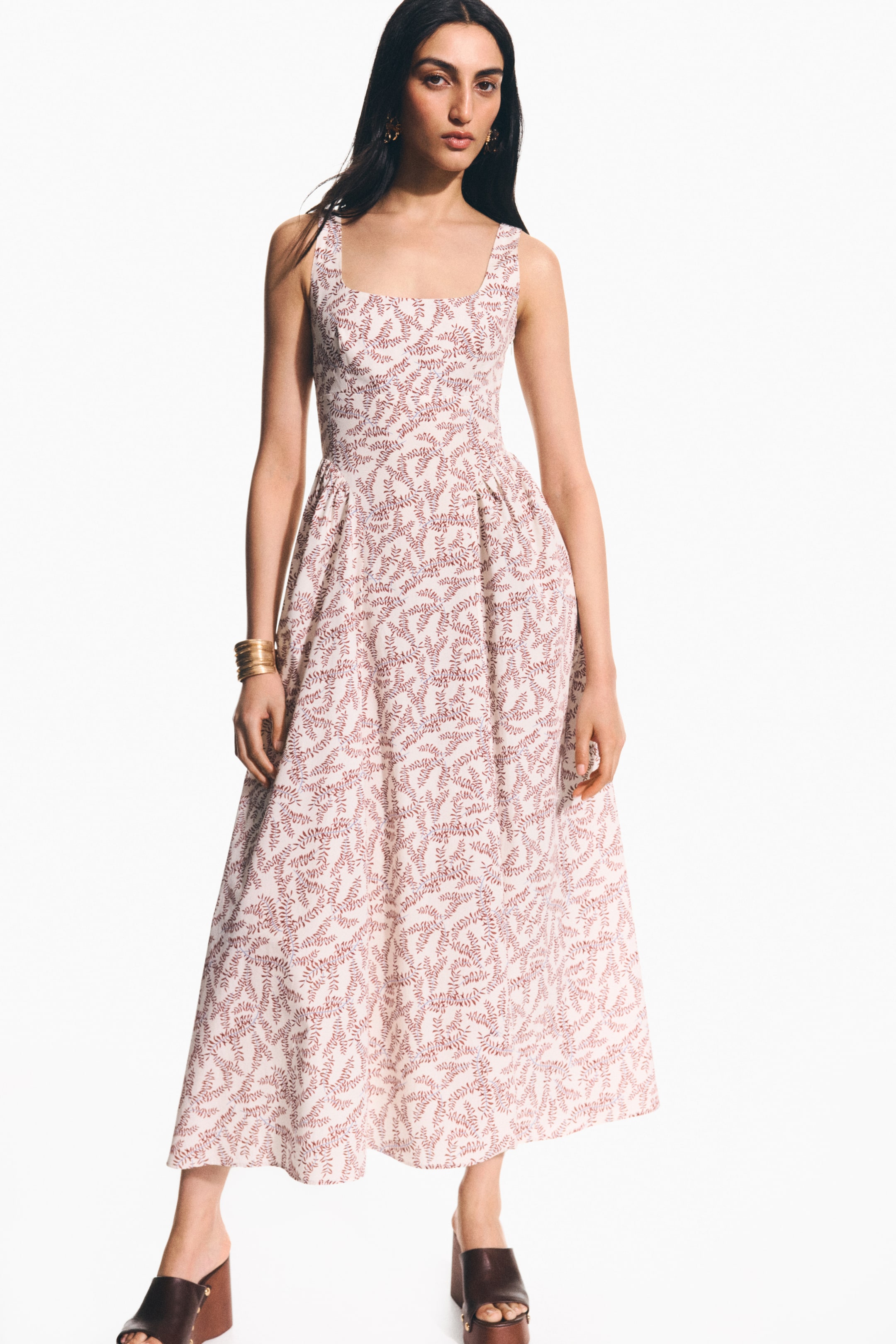 View larger image: A calf-length dress in a white cotton weave with a reddish-brown leaf pattern, designed with a square neckline, wide shoulder straps, a fitted bodice, and a flared skirt that creates volume with gathers, is being worn by the model. The model is also wearing golden bangles on her right wrist and dark brown platform mules.