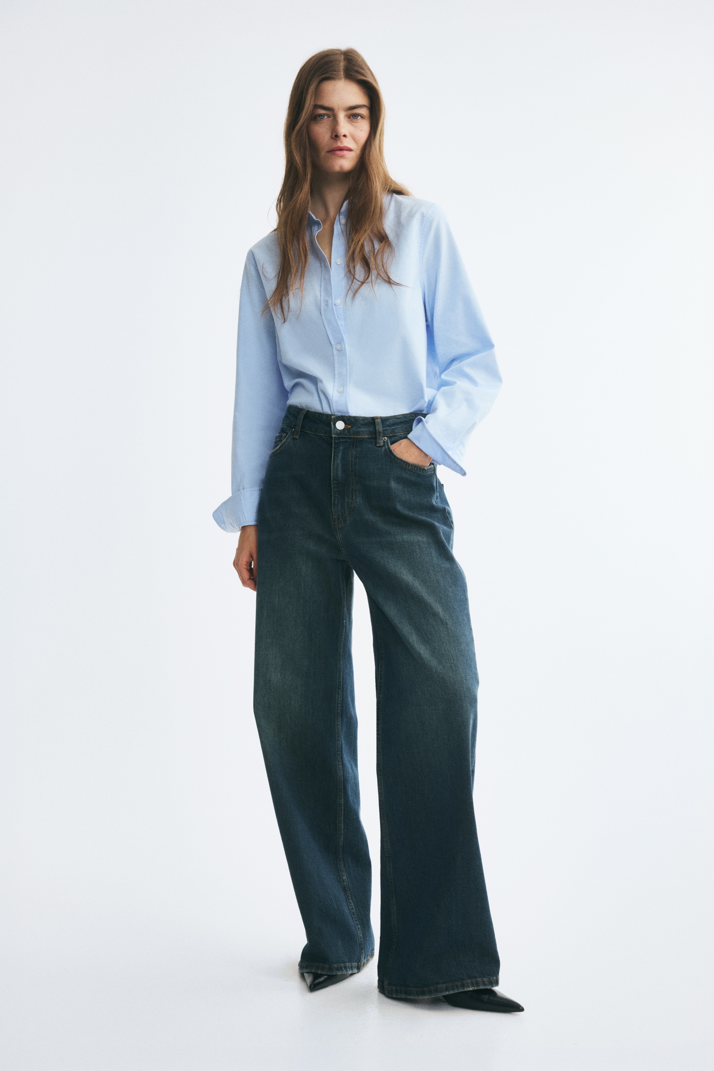 A pair of dark blue denim jeans is hanging vertically in a front view, showcasing a loose, straight-leg fit with a faded, worn-in wash along the thighs. The design features a regular waist with belt loops, a button fastening, and classic five-pocket styling with slightly distressed pocket edges.
