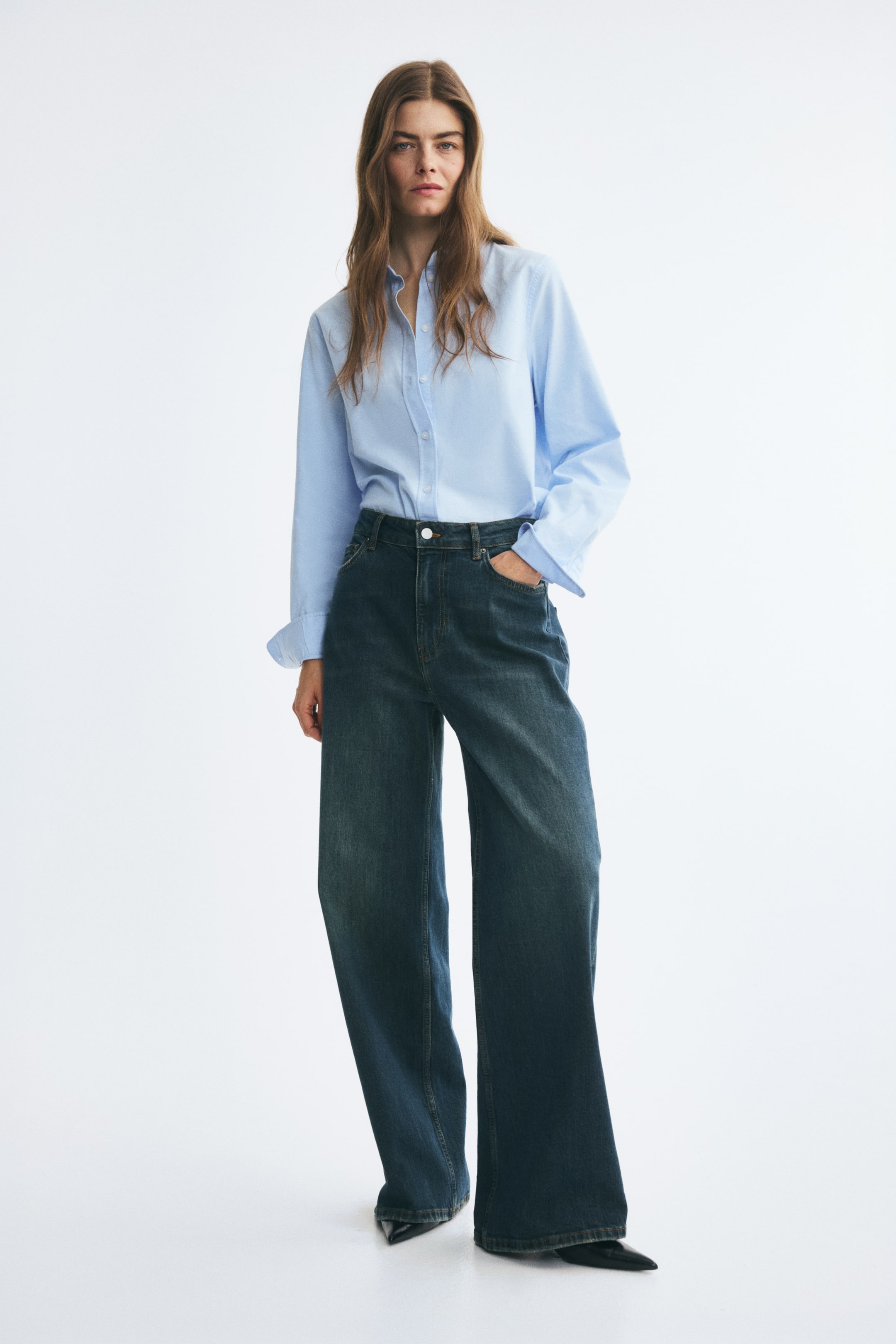 Ver imagem maior: Jeans de ganga azul escura com lavagem escura, cintura regular e corte largo e direito, usados pela modelo. A modelo está de pé com uma mão no bolso da frente, estilizando as jeans com uma camisa azul-clara de mangas compridas, metida por dentro, e sapatos pretos de bico fino.