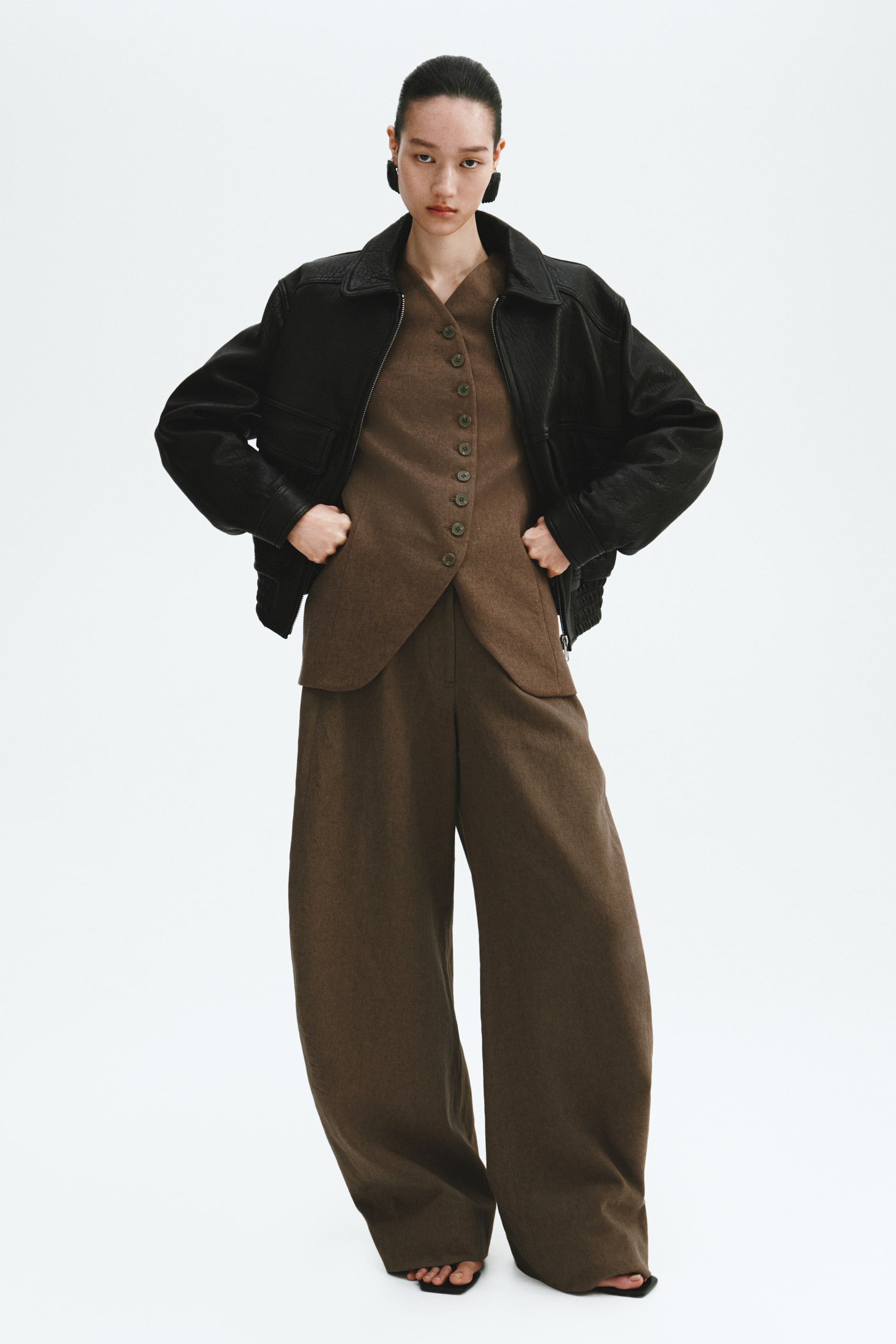 View larger image: A black textured bomber jacket with a wide collar is worn over a brown V-neck button-front vest and matching full-length wide-leg trousers. The model has hands on hips.